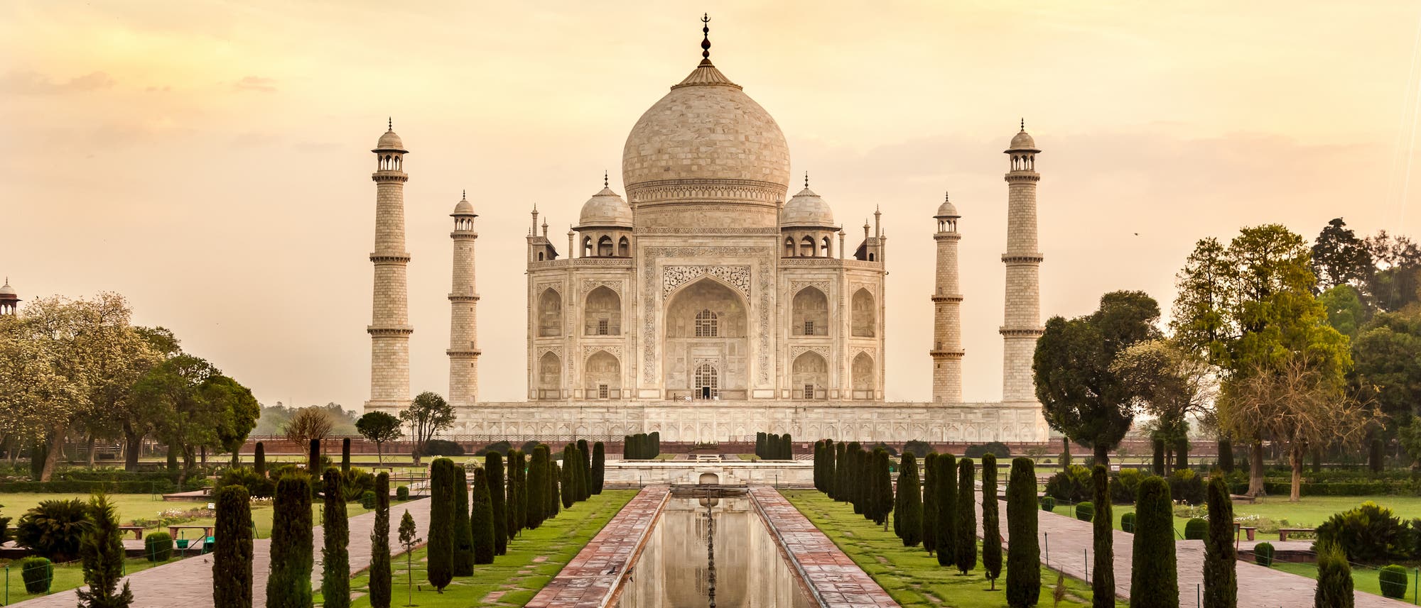 Taj Mahal, ein großes, weißes Marmorgebäude mit einer zentralen Kuppel und vier Minaretten, umgeben von gepflegten Gärten und einem reflektierenden Wasserbecken im Vordergrund. Der Himmel ist leicht bewölkt und in warmen Tönen gehalten.
