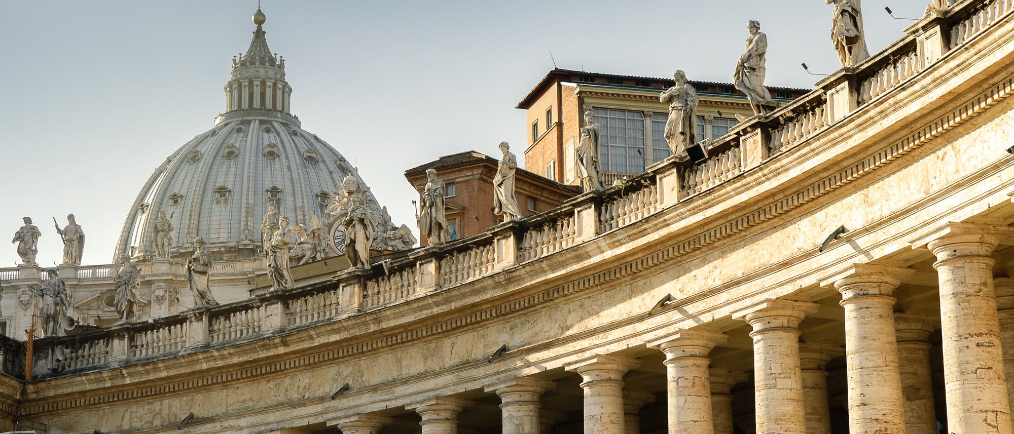 Eine Ansicht der Kolonnaden des Petersplatzes im Vatikan, die von zahlreichen Statuen gesäumt sind. Im Hintergrund ist die Kuppel des Petersdoms sichtbar. Die Architektur zeigt klassische Säulen und barocke Details. Der Himmel ist klar und blau. Keine Personen sind im Bild zu sehen.