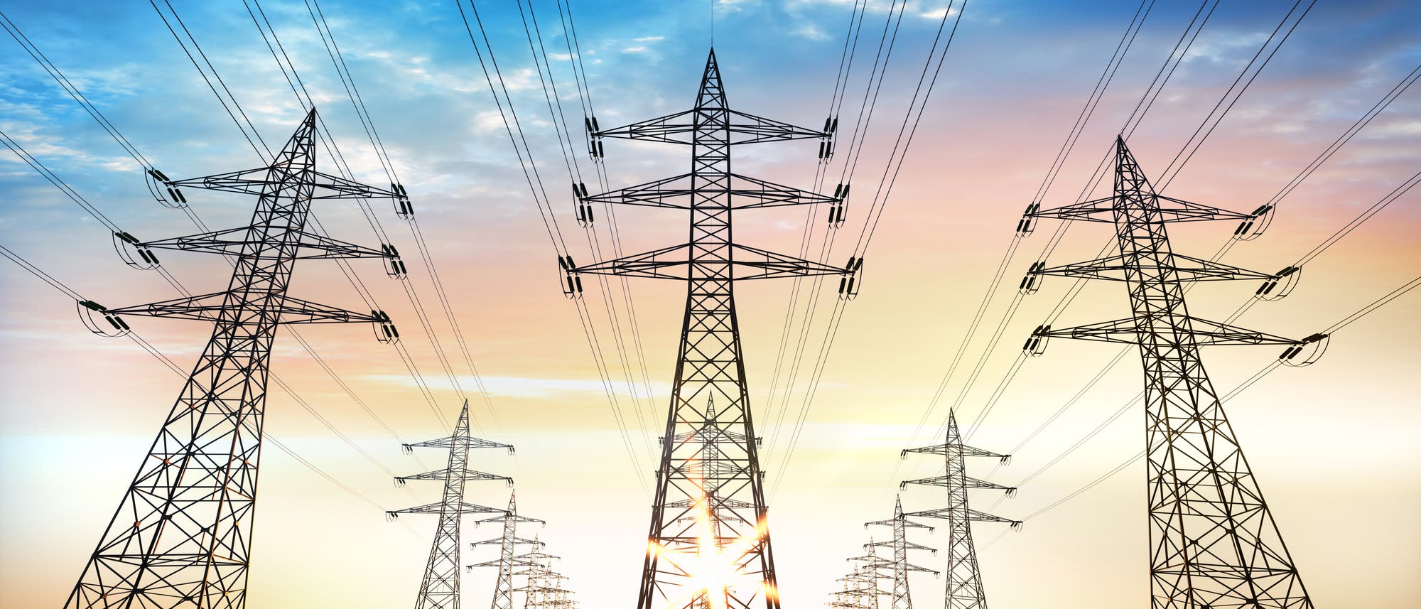 Mehrere Strommasten stehen in einer Reihe und tragen elektrische Leitungen, die sich in die Ferne erstrecken. Der Himmel im Hintergrund zeigt einen Farbverlauf von Blau zu Orange, was auf einen Sonnenauf- oder -untergang hindeutet. Die Szene vermittelt ein Gefühl von Weite und Energieinfrastruktur.