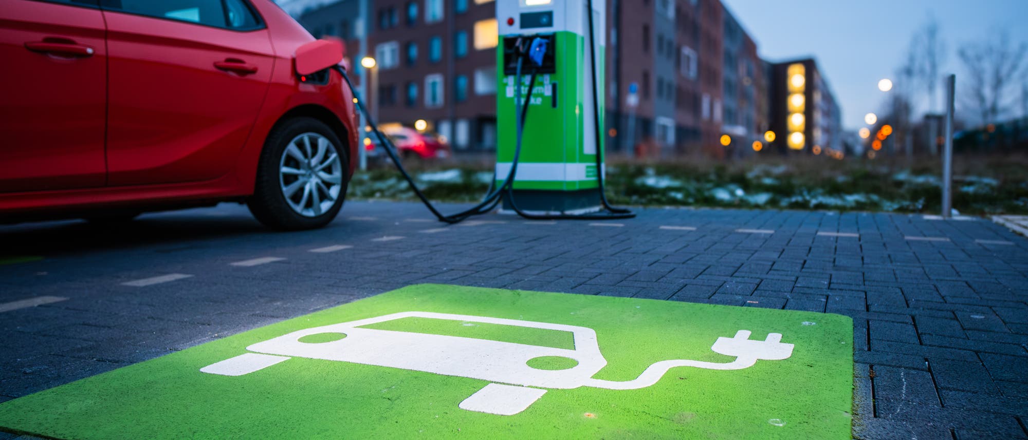 Ein rotes Elektroauto ist an eine grüne Ladestation angeschlossen, die sich auf einem Parkplatz in einer städtischen Umgebung befindet. Der Boden ist mit einem grünen Symbol für Elektrofahrzeuge markiert. Im Hintergrund sind moderne Wohngebäude und Straßenlaternen zu sehen. Die Szene vermittelt eine nachhaltige und umweltfreundliche Mobilität.