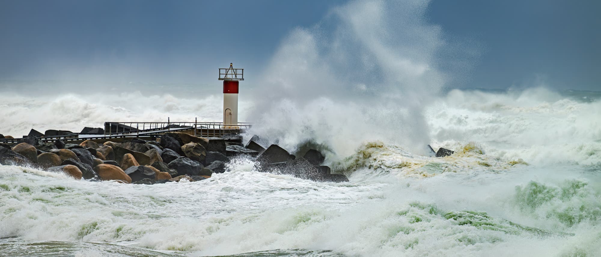 Stürmische Meereswellen schlagen gegen einen Leuchtturm auf einer Mole. Der Himmel ist dunkel und bewölkt, während Gischt hoch in die Luft spritzt. Eine Möwe fliegt in der Nähe, während die Wellen tosen. Die Szene vermittelt die Kraft und Dynamik des Meeres bei rauem Wetter. Stürmische Meereswellen schlagen gegen einen Leuchtturm auf einer Mole. Der Himmel ist dunkel und bewölkt, während Gischt hoch in die Luft spritzt. Eine Möwe fliegt in der Nähe, während die Wellen tosen. Die Szene vermittelt die Kraft und Dynamik des Meeres bei rauem Wetter.