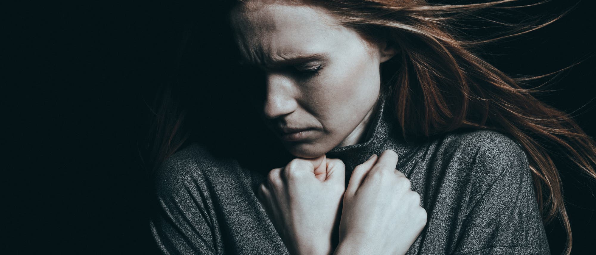 Eine Frau in einem dunklen Raum hält ihre Hände vor der Brust und blickt nach unten. Der Gesichtsausdruck wirkt nachdenklich oder besorgt. 