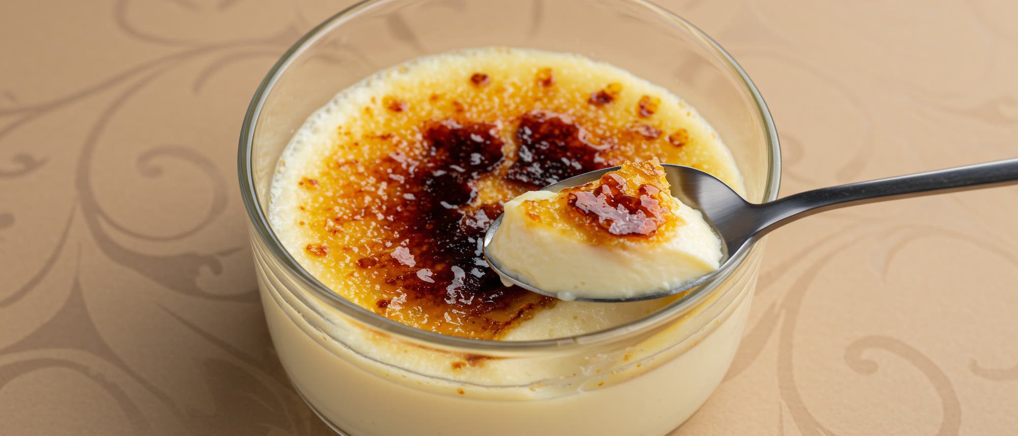 Ein Glas mit Crème Brûlée, einer französischen Nachspeise, auf einem dekorativen Tisch. Die karamellisierte Zuckerschicht ist knusprig gebräunt. Ein Löffel hebt eine Portion der cremigen Vanillepuddingmasse aus dem Glas. Ein Glas mit Crème Brûlée, einer französischen Nachspeise, auf einem dekorativen Tisch. Die karamellisierte Zuckerschicht ist knusprig gebräunt. Ein Löffel hebt eine Portion der cremigen Vanillepuddingmasse aus dem Glas.