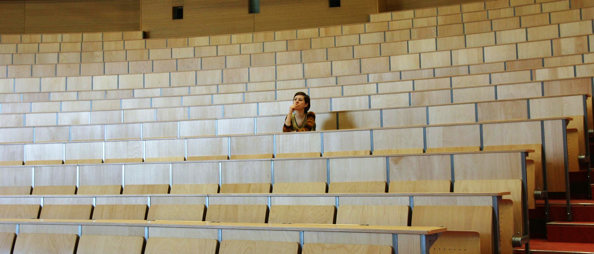 Eine Person sitzt allein in einem großen, leeren Hörsaal mit hölzernen Sitzreihen. Die Person scheint nachdenklich zu sein und blickt nach vorne. Der Raum ist gut beleuchtet und die Wände sind aus Holz. Eine Person sitzt allein in einem großen, leeren Hörsaal mit hölzernen Sitzreihen. Die Person scheint nachdenklich zu sein und blickt nach vorne. Der Raum ist gut beleuchtet und die Wände sind aus Holz.