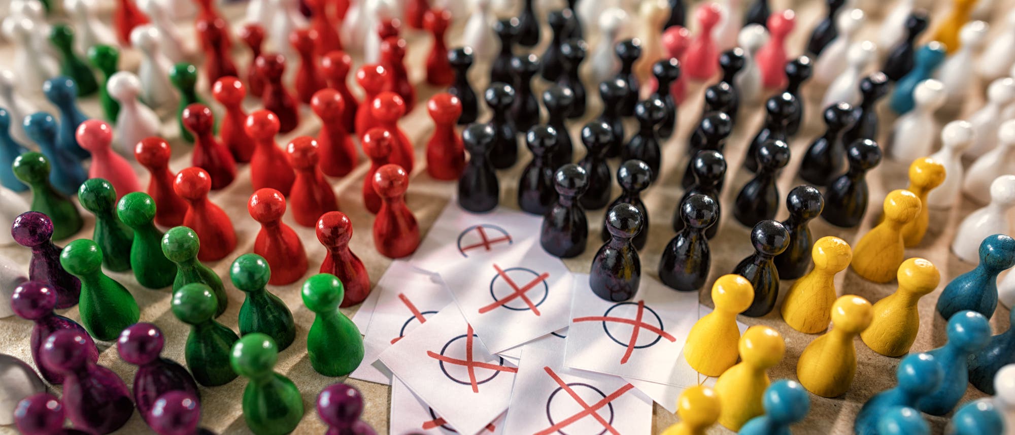 Eine gro&szlig;e Anzahl bunter Spielfiguren ist in Gruppen aufgestellt, die verschiedene Farben wie Rot, Gr&uuml;n, Blau, Schwarz, Wei&szlig;, Gelb und Lila repr&auml;sentieren. In der Mitte liegen mehrere wei&szlig;e Karten mit roten Kreuzen darauf. Die Anordnung der Figuren und Karten k&ouml;nnte symbolisch f&uuml;r Wahlen oder Entscheidungsprozesse stehen.