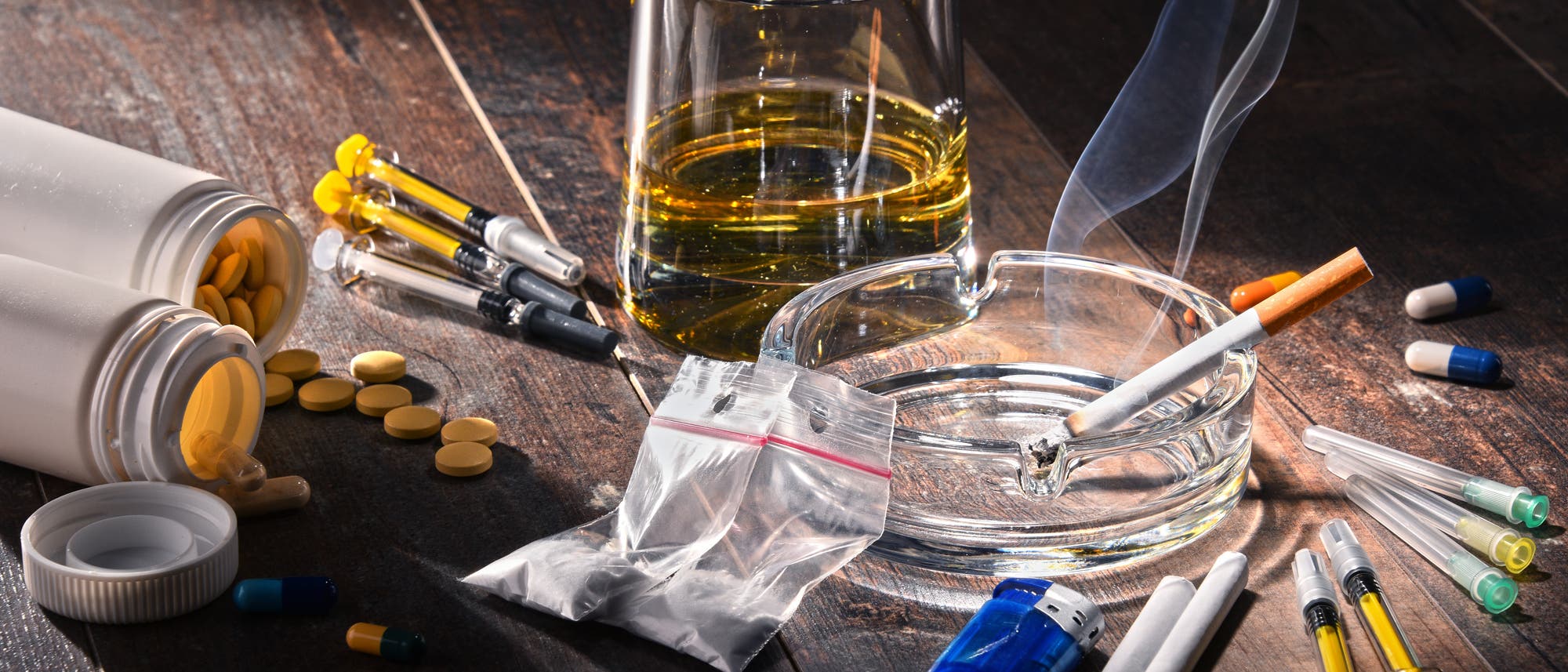 Auf einem Holztisch liegen verschiedene Drogenutensilien verstreut. Zu sehen sind umgefallene Pillenfläschchen mit Tabletten, Spritzen, ein Glas mit einer alkoholischen Flüssigkeit, ein Aschenbecher mit einer brennenden Zigarette und ein kleines Plastiktütchen. Auf einem Holztisch liegen verschiedene Drogenutensilien verstreut. Zu sehen sind umgefallene Pillenfläschchen mit Tabletten, Spritzen, ein Glas mit einer alkoholischen Flüssigkeit, ein Aschenbecher mit einer brennenden Zigarette und ein kleines Plastiktütchen.