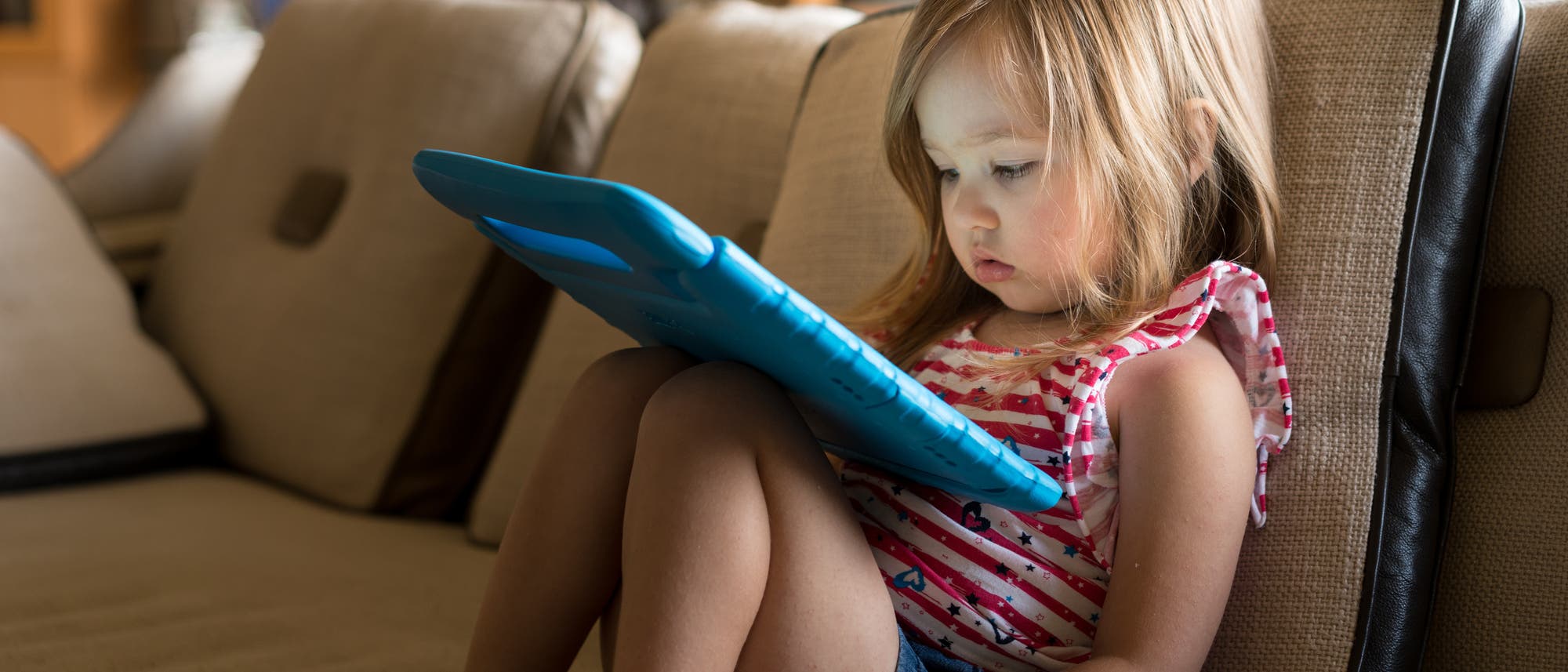 Ein kleines Mädchen sitzt auf dem Sofa und schaut in eine Art Tablet 