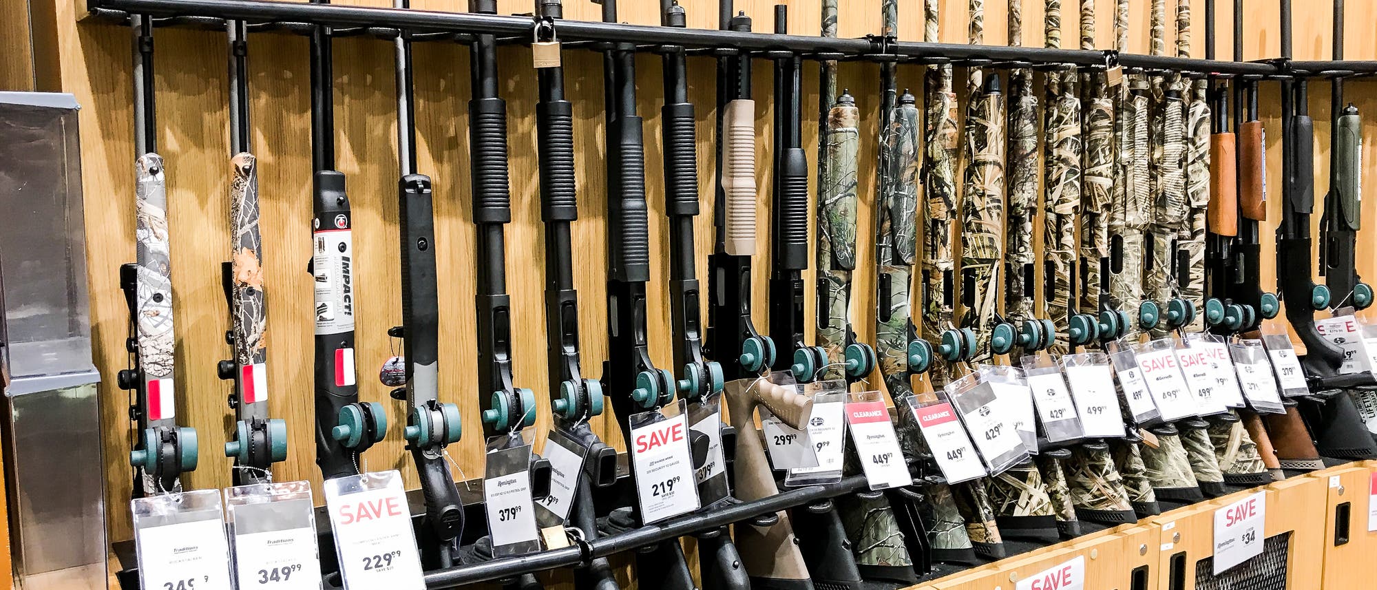 Eine Reihe von Gewehren ist in einem Gesch&auml;ft auf einem Holzregal ausgestellt. Jedes Gewehr ist mit einem Preisschild versehen, das Rabatte anzeigt. Die Gewehre sind in verschiedenen Designs und Mustern gehalten. Im Vordergrund sind mehrere Schilder mit der Aufschrift "SALE" zu sehen, die auf reduzierte Preise hinweisen.