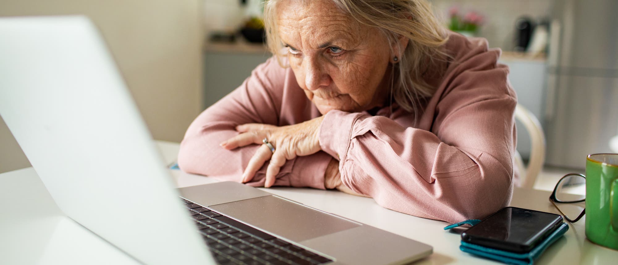 Eine Person sitzt an einem Tisch und schaut konzentriert auf einen Laptop-Bildschirm. Neben dem Laptop liegen ein Smartphone und eine Brille. Im Hintergrund ist eine K&uuml;che zu sehen. Die Person tr&auml;gt ein rosa Hemd und st&uuml;tzt den Kopf auf die H&auml;nde.