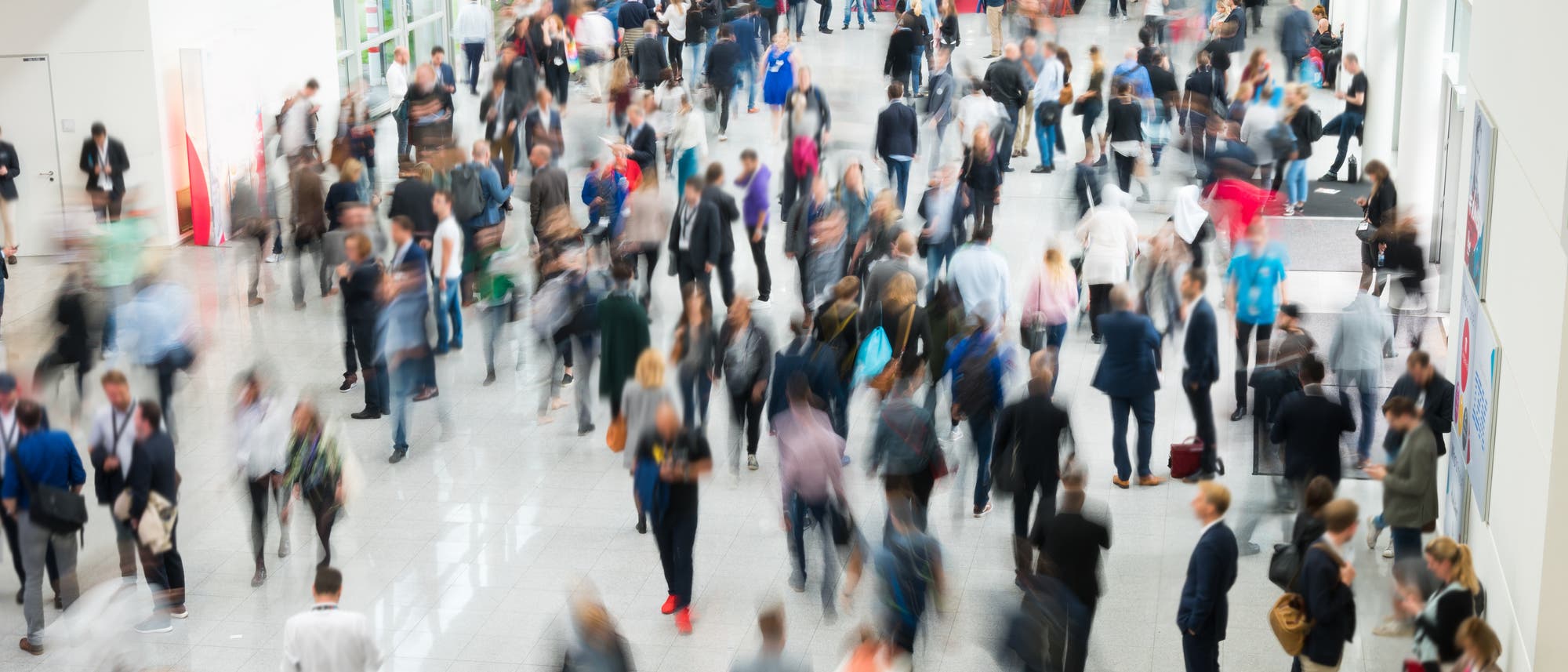 Eine gro&szlig;e Menschenmenge bewegt sich durch eine helle, ger&auml;umige Halle. Die Personen sind unscharf dargestellt, was auf Bewegung hinweist. Einige tragen formelle Kleidung, w&auml;hrend andere l&auml;ssiger gekleidet sind. Die Umgebung wirkt gesch&auml;ftig und lebendig, typisch f&uuml;r eine Messe oder Konferenz. Im Hintergrund sind wei&szlig;e W&auml;nde und gro&szlig;e Fenster zu sehen, die viel Licht hereinlassen.