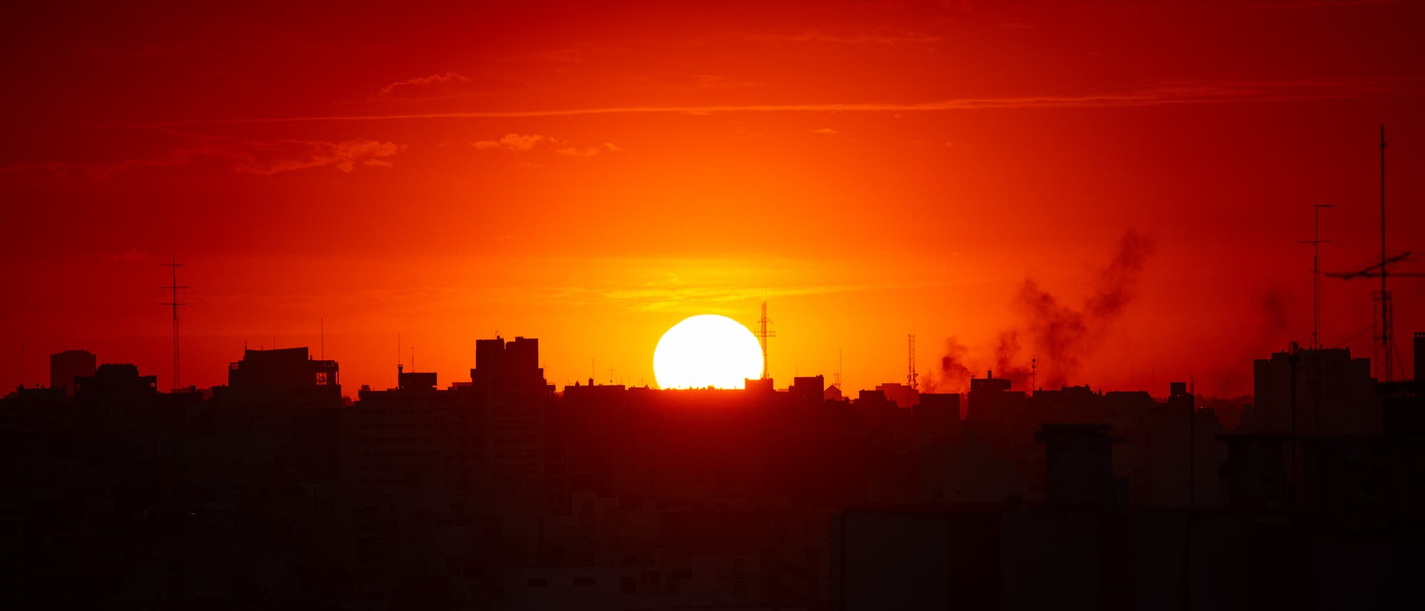 Sonnenuntergang über einer Stadtlandschaft. Die untergehende Sonne taucht den Himmel in ein intensives Rot-Orange, während die Silhouetten von Gebäuden und Antennen im Vordergrund sichtbar sind. Rauchwolken steigen von einigen Gebäuden auf, was der Szene eine dynamische Note verleiht. Sonnenuntergang über einer Stadtlandschaft. Die untergehende Sonne taucht den Himmel in ein intensives Rot-Orange, während die Silhouetten von Gebäuden und Antennen im Vordergrund sichtbar sind. Rauchwolken steigen von einigen Gebäuden auf, was der Szene eine dynamische Note verleiht.