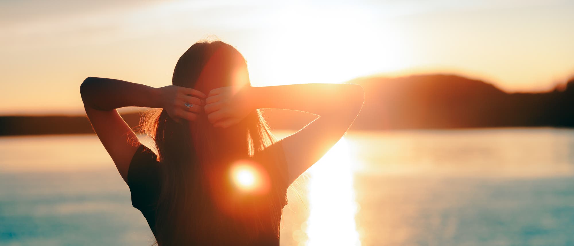 Man sieht eine Frau mit verschränkten Armen von hinten, sie genießt den Sonnenuntergang am See.