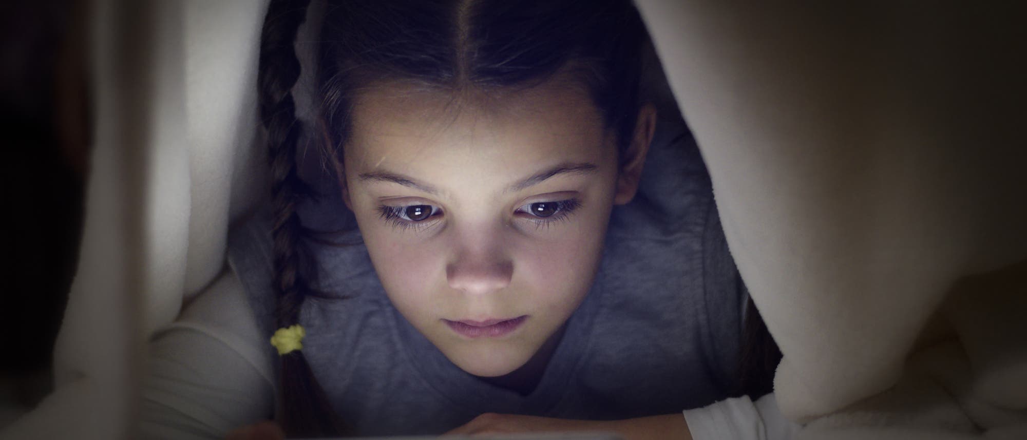 Ein junges Mädchen liegt unter einer Bettdecke und guckt konzentriert auf ein Bildschirmgerät.