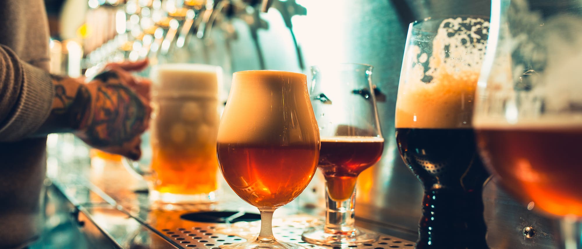 Eine Reihe von Biergläsern mit verschiedenen Biersorten steht auf einer Theke in einer Bar. Im Hintergrund ist eine Person zu sehen, die ein weiteres Glas unter einen Zapfhahn hält. Die Gläser sind unterschiedlich geformt und gefüllt, einige mit Schaumkronen. Die Szene ist in warmem Licht gehalten, das die Farben der Biere betont.
