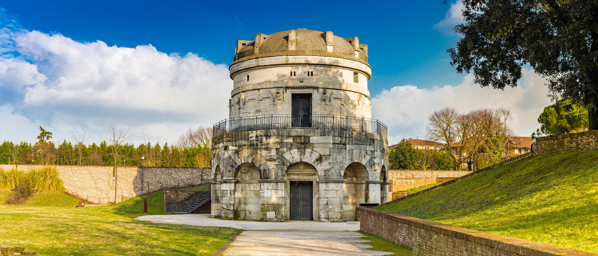 Ein historisches, rundes Steingebäude mit einer Kuppel, umgeben von einer grünen Wiese und Bäumen. Der Himmel ist blau mit einigen Wolken. Das Gebäude hat zwei Ebenen mit Bögen und eine umlaufende Balustrade. Im Hintergrund sind eine Mauer und weitere Bäume zu sehen. Ein historisches, rundes Steingebäude mit einer Kuppel, umgeben von einer grünen Wiese und Bäumen. Der Himmel ist blau mit einigen Wolken. Das Gebäude hat zwei Ebenen mit Bögen und eine umlaufende Balustrade. Im Hintergrund sind eine Mauer und weitere Bäume zu sehen.