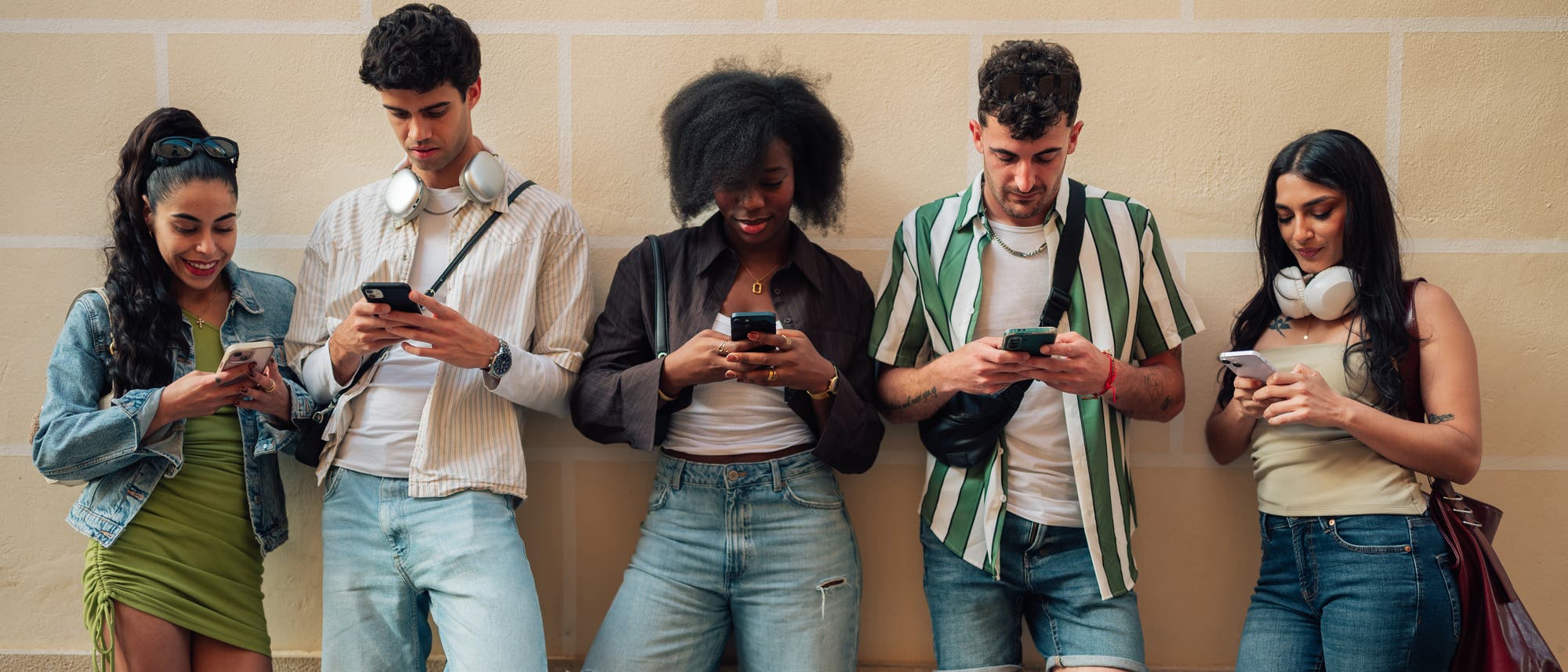 Eine Gruppe von fünf jungen Erwachsenen steht vor einer beigen Wand und schaut auf ihre Smartphones. Sie tragen lässige Kleidung, darunter Jeans und T-Shirts, und einige haben Kopfhörer um den Hals. Die Szene vermittelt eine entspannte Atmosphäre und zeigt den alltäglichen Gebrauch von Technologie. Eine Gruppe von fünf jungen Erwachsenen steht vor einer beigen Wand und schaut auf ihre Smartphones. Sie tragen lässige Kleidung, darunter Jeans und T-Shirts, und einige haben Kopfhörer um den Hals. Die Szene vermittelt eine entspannte Atmosphäre und zeigt den alltäglichen Gebrauch von Technologie.
