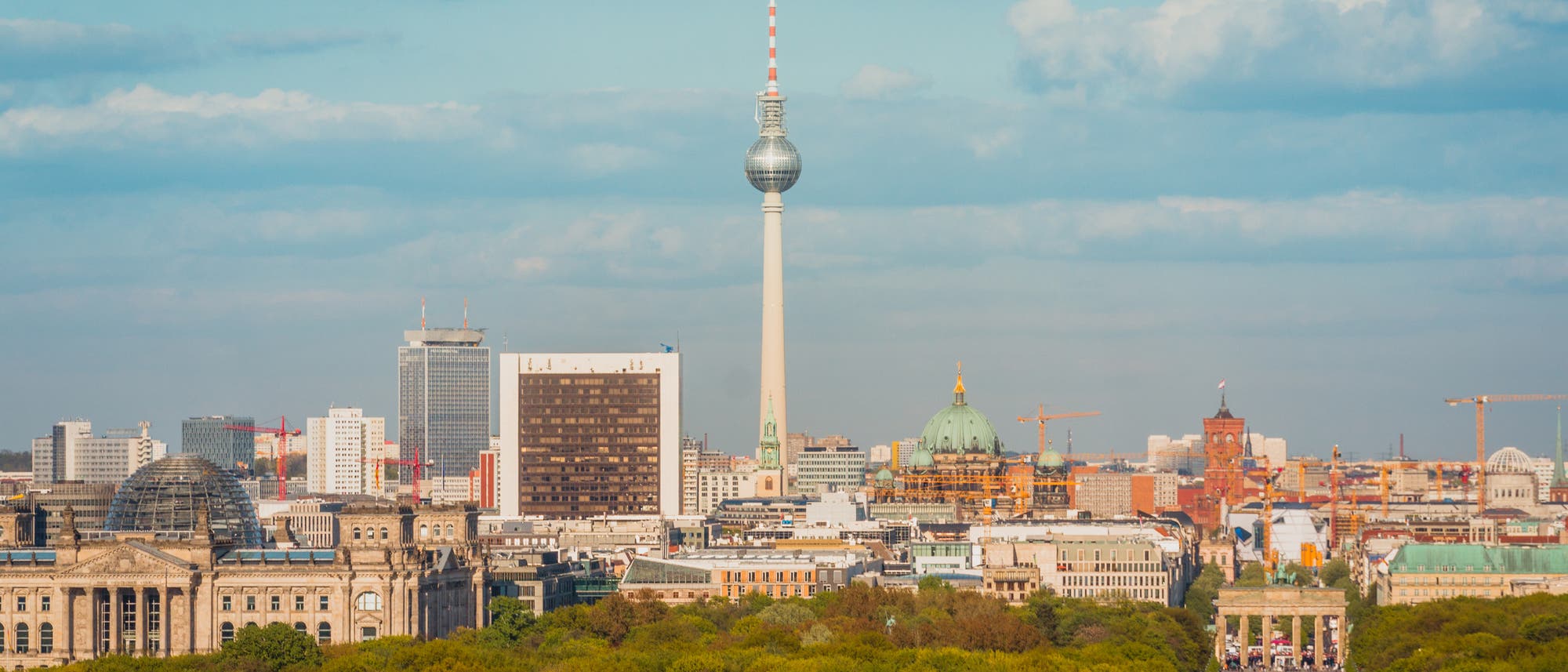 Stadtansicht von Berlin mit dem Fernsehturm im Zentrum, umgeben von modernen und historischen Gebäuden. Im Vordergrund erstreckt sich der Tiergarten, während das Brandenburger Tor rechts sichtbar ist. Der Himmel ist leicht bewölkt. Stadtansicht von Berlin mit dem Fernsehturm im Zentrum, umgeben von modernen und historischen Gebäuden. Im Vordergrund erstreckt sich der Tiergarten, während das Brandenburger Tor rechts sichtbar ist. Der Himmel ist leicht bewölkt.