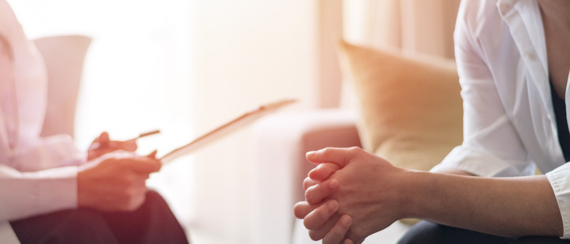 Zwei Personen sitzen in einem hellen Raum. Die Person links hält einen Stift und ein Klemmbrett, während die Person rechts die Hände gefaltet hat. Beide tragen weiße Hemden. Die Szene deutet auf ein Gespräch oder eine Beratung hin. Im Hintergrund sind verschwommene Möbel zu sehen. Zwei Personen sitzen in einem hellen Raum. Die Person links hält einen Stift und ein Klemmbrett, während die Person rechts die Hände gefaltet hat. Beide tragen weiße Hemden. Die Szene deutet auf ein Gespräch oder eine Beratung hin. Im Hintergrund sind verschwommene Möbel zu sehen.