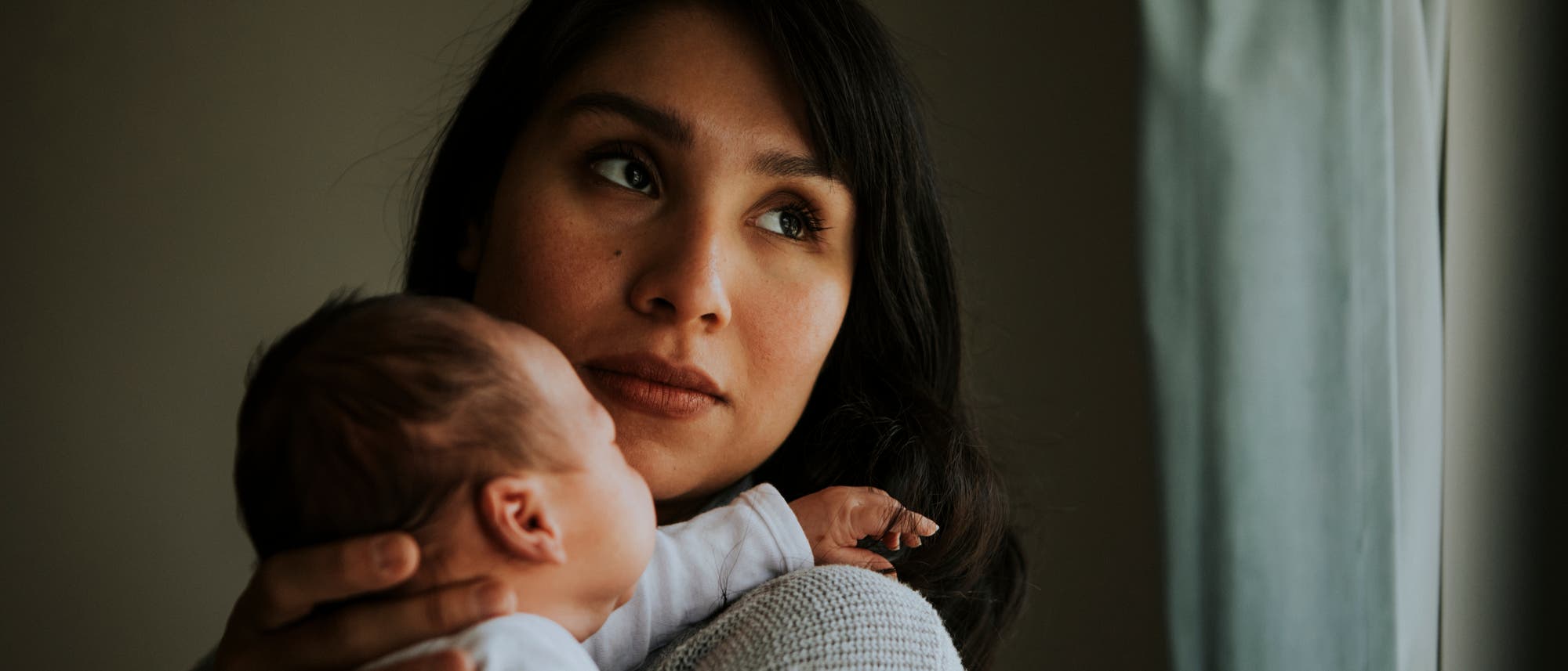 Eine Frau hält ein Baby auf dem Arm und schaut nachdenklich aus einem Fenster. Die Szene ist in warmem Licht gehalten, das durch das Fenster fällt. Eine Frau hält ein Baby auf dem Arm und schaut nachdenklich aus einem Fenster. Die Szene ist in warmem Licht gehalten, das durch das Fenster fällt.