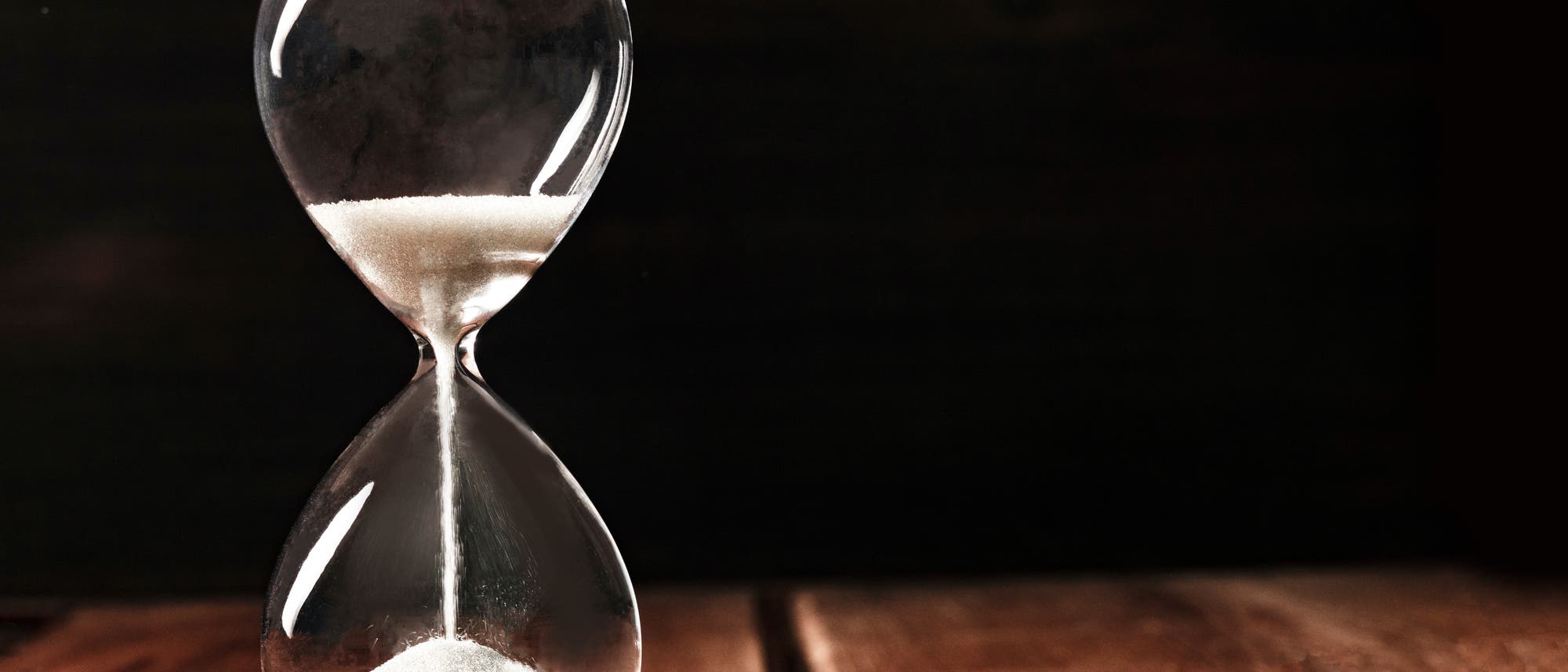 Eine Sanduhr aus Glas vor einem schwarzen Hintergrund symbolisiert Zeitdruck und Arbeitsbelastung Eine Sanduhr aus Glas vor einem schwarzen Hintergrund symbolisiert Zeitdruck und Arbeitsbelastung