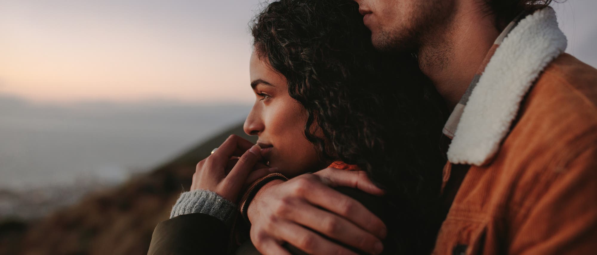 Ein Paar schaut in der Natur Richtung Horizont. Der Mann hält seine Partnerin fest in den Armen.