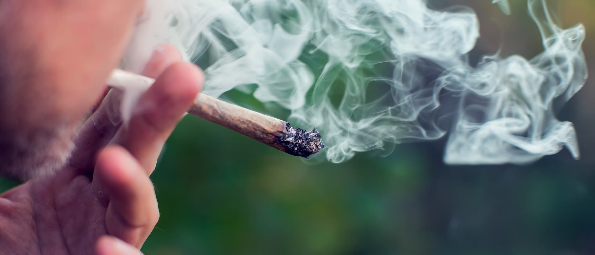 Eine Person h&auml;lt einen brennenden Joint in der Hand, w&auml;hrend Rauch in die Luft aufsteigt. Der Hintergrund ist unscharf und gr&uuml;nlich, was auf eine Au&szlig;enumgebung hindeutet. Der Fokus liegt auf dem Joint und dem Rauch, der sich in geschwungenen Mustern ausbreitet.
