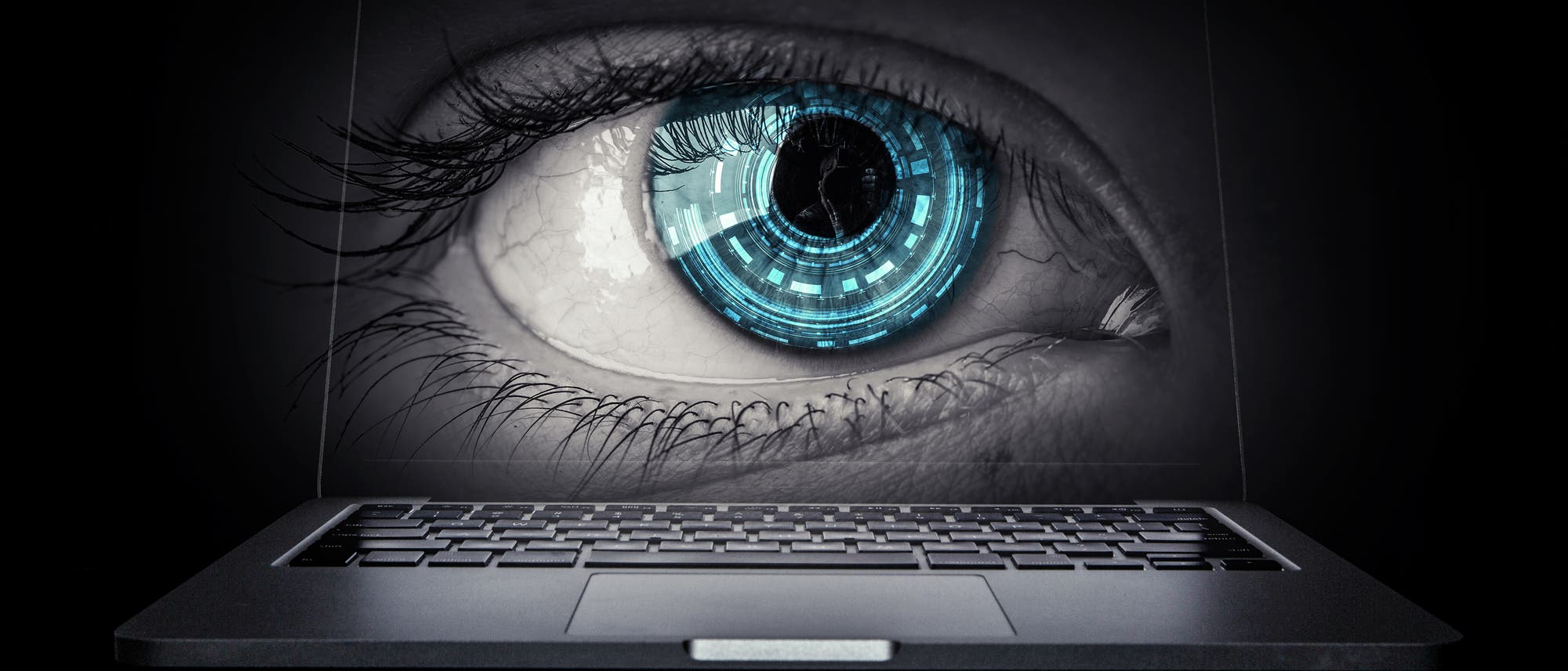 Ein Laptop mit einem gro&szlig;en, digitalen Auge auf dem Bildschirm vor einem sehr dunklen Hintergrund. 