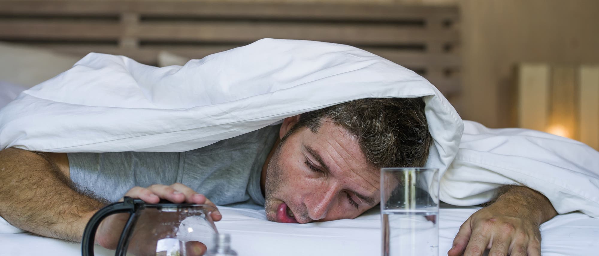 Ein verkaterter Mann guckt unter einer Bettdecke hervor.