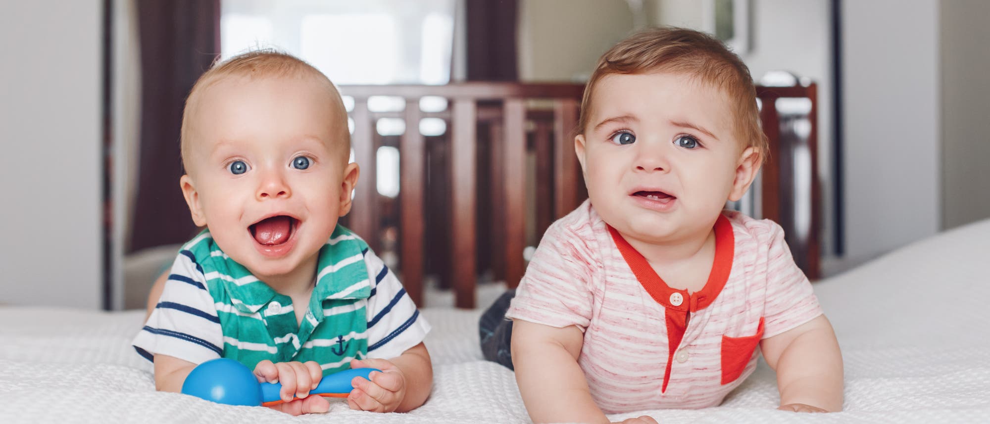 Zwei Babys liegen auf einem Bett. Das Baby links trägt ein gestreiftes Shirt, lächelt und hält ein blaues Spielzeug. Das Baby rechts trägt ein gestreiftes Shirt mit rotem Kragen und sieht besorgt aus. Im Hintergrund sind ein Kinderbett und ein Fenster zu sehen.
