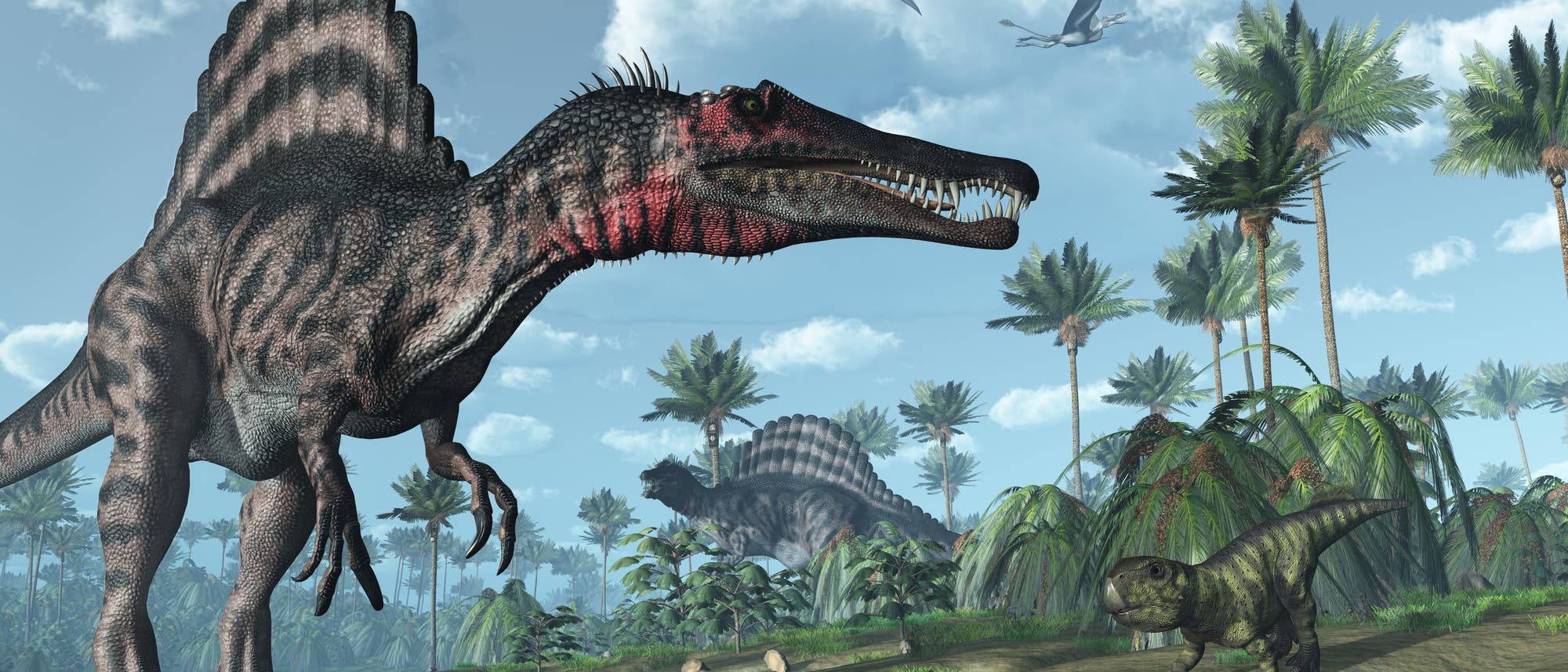 Eine 3D-Darstellung einer prähistorischen Landschaft mit mehreren Dinosauriern. Im Vordergrund steht ein großer Dinosaurier mit einem segelartigen Rücken, der am Ufer eines Gewässers steht. Im Hintergrund sind weitere Dinosaurier und üppige Vegetation mit Palmen zu sehen. Am Himmel fliegen zwei Flugsaurier. Die Szene vermittelt eine lebendige Darstellung der Urzeit.