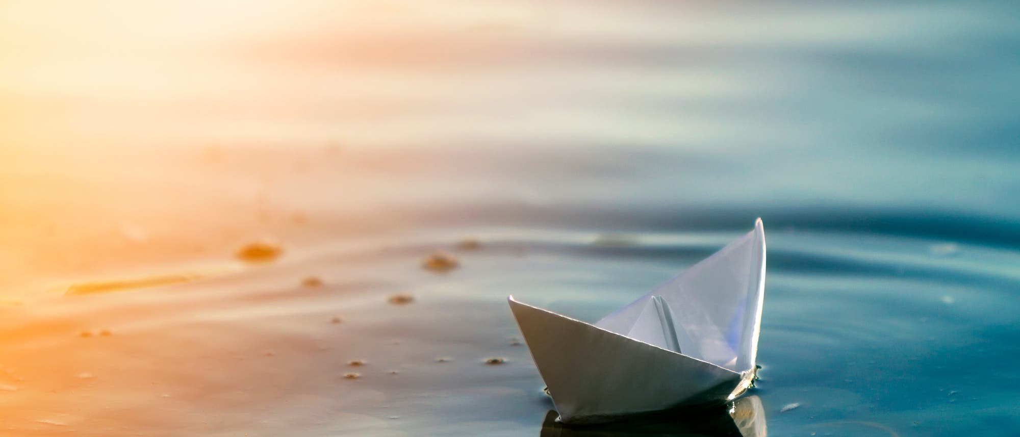 Ein kleines Papierboot schwimmt auf ruhigem Wasser, das in warmen Sonnenlichtt&ouml;nen leuchtet. Die Oberfl&auml;che des Wassers spiegelt das Boot wider und erzeugt eine friedliche, vertr&auml;umte Atmosph&auml;re.