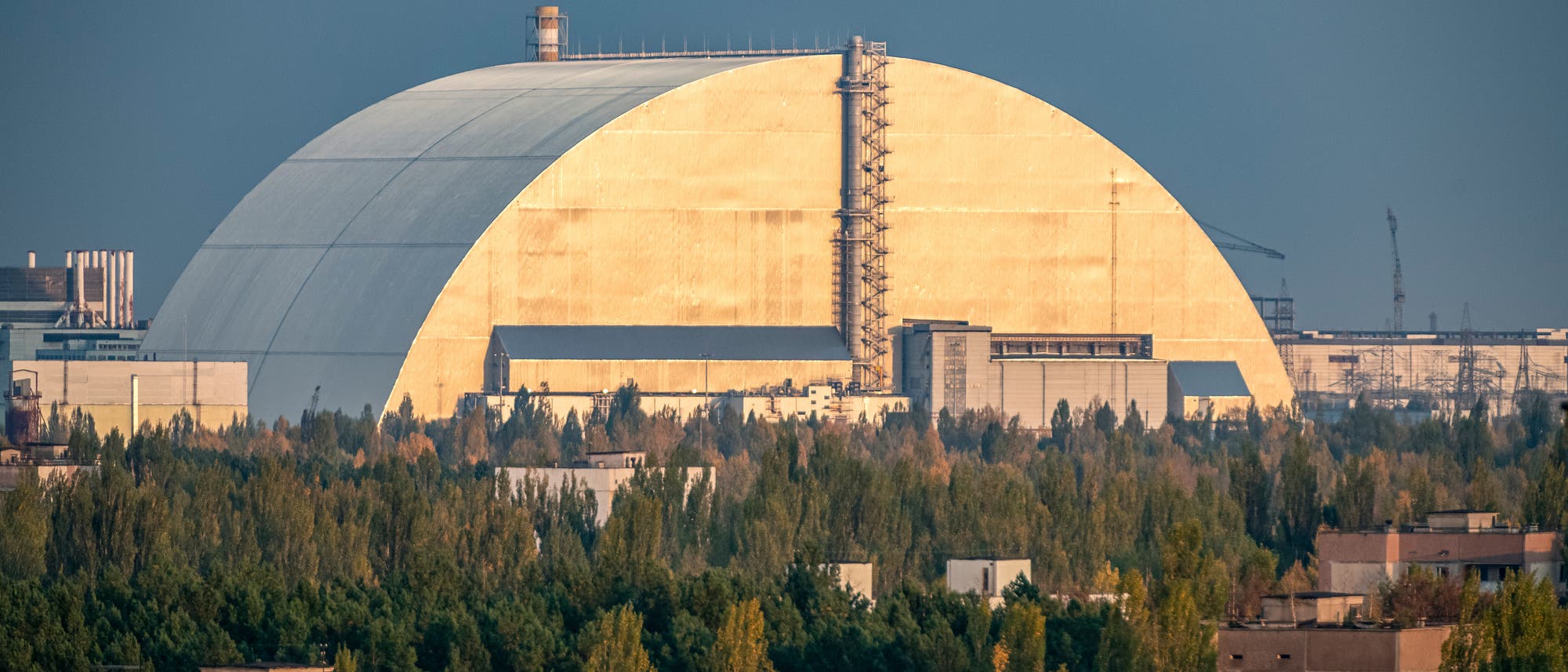 Eine gro&szlig;e, gew&ouml;lbte Struktur aus Metall, die von B&auml;umen und Geb&auml;uden umgeben ist. Die Struktur hat eine helle, reflektierende Oberfl&auml;che und ist von einem klaren Himmel umgeben. Im Vordergrund sind mehrere flache Geb&auml;ude und eine dichte Baumreihe zu sehen.
