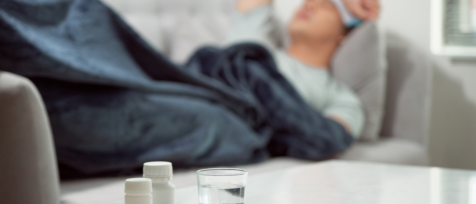 Eine Person liegt auf einem Sofa, zugedeckt mit einer dunklen Decke, und h&auml;lt sich den Kopf. Im Vordergrund auf einem Tisch stehen zwei Medikamentenflaschen, ein Glas Wasser, ein Thermometer und eine Blisterpackung mit Tabletten. Die Szene deutet auf eine kranke Person hin, die sich ausruht.