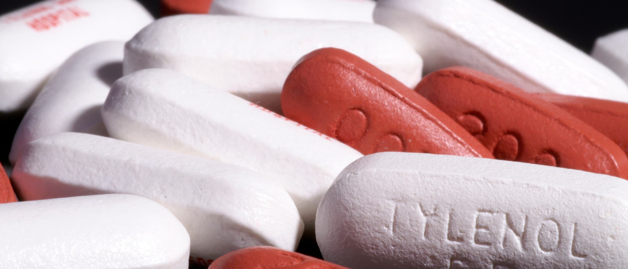 Eine Nahaufnahme von mehreren weißen und roten Tabletten auf schwarzem Hintergrund. Auf einer der weißen Tabletten ist der Text "TYLENOL ER" eingeprägt. Die roten Tabletten haben die Zahl "500" eingeprägt. Das Bild zeigt eine Mischung von Medikamenten in Kapselform.