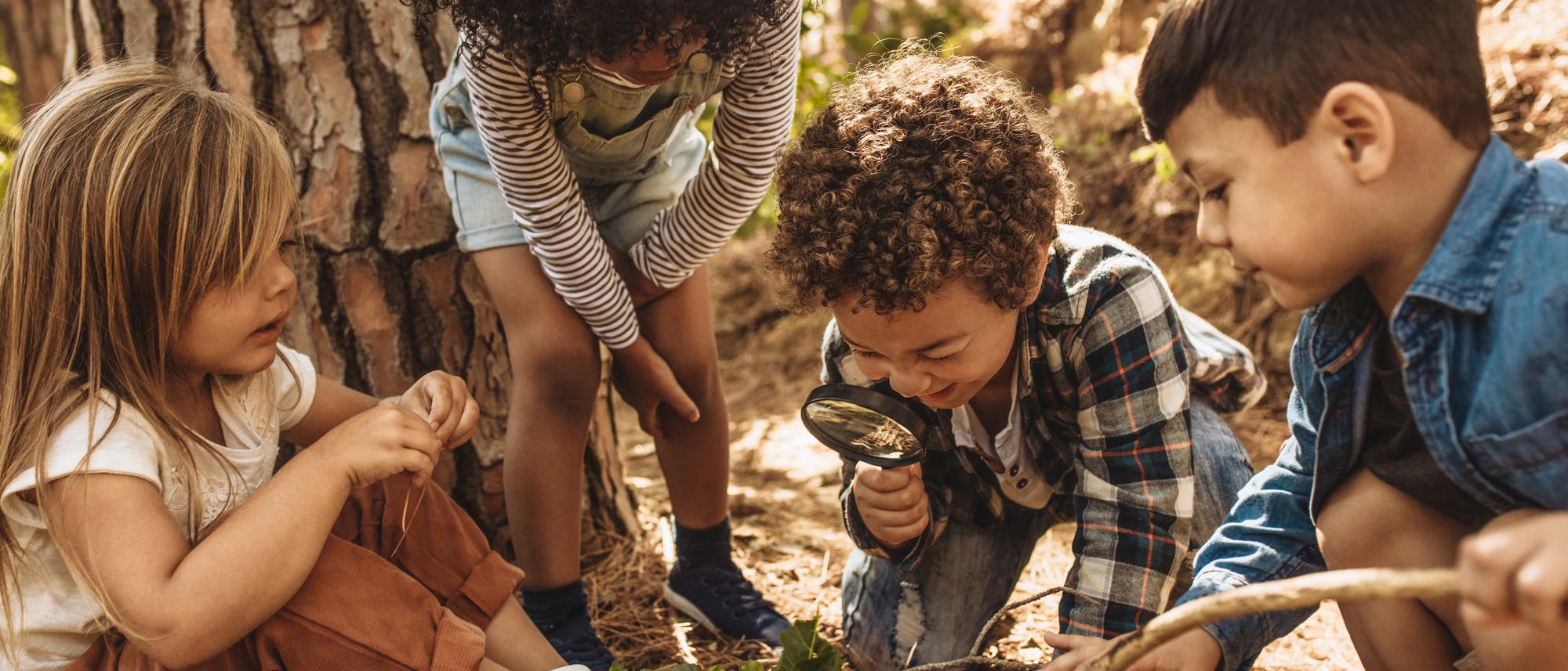 Eine Gruppe von vier Kindern spielt im Freien im Wald. Ein Kind h&auml;lt eine Lupe und untersucht den Boden, w&auml;hrend die anderen neugierig zuschauen. Die Szene ist von B&auml;umen und nat&uuml;rlichem Licht umgeben, was eine Atmosph&auml;re von Entdeckung und Abenteuer schafft.