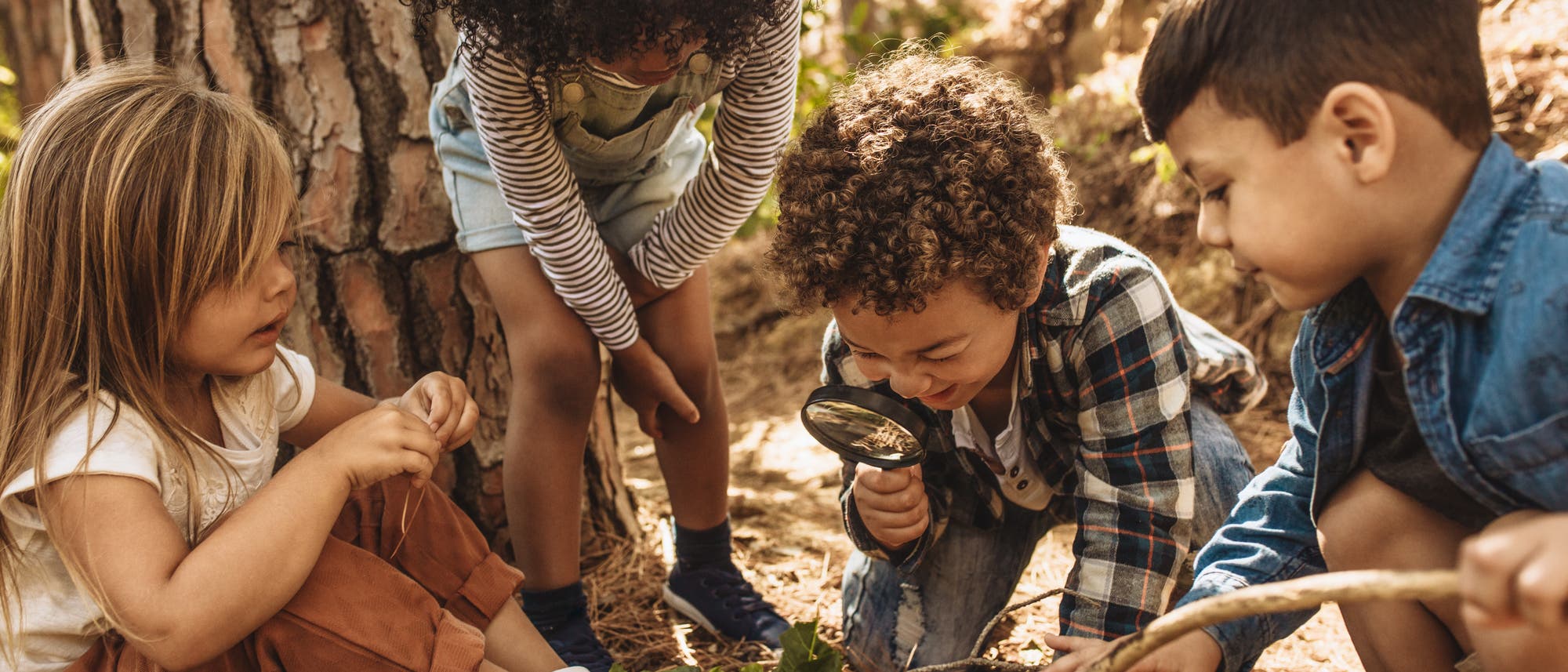Eine Gruppe von vier Kindern spielt im Freien im Wald. Ein Kind h&auml;lt eine Lupe und untersucht den Boden, w&auml;hrend die anderen neugierig zuschauen. Die Szene ist von B&auml;umen und nat&uuml;rlichem Licht umgeben, was eine Atmosph&auml;re von Entdeckung und Abenteuer schafft.