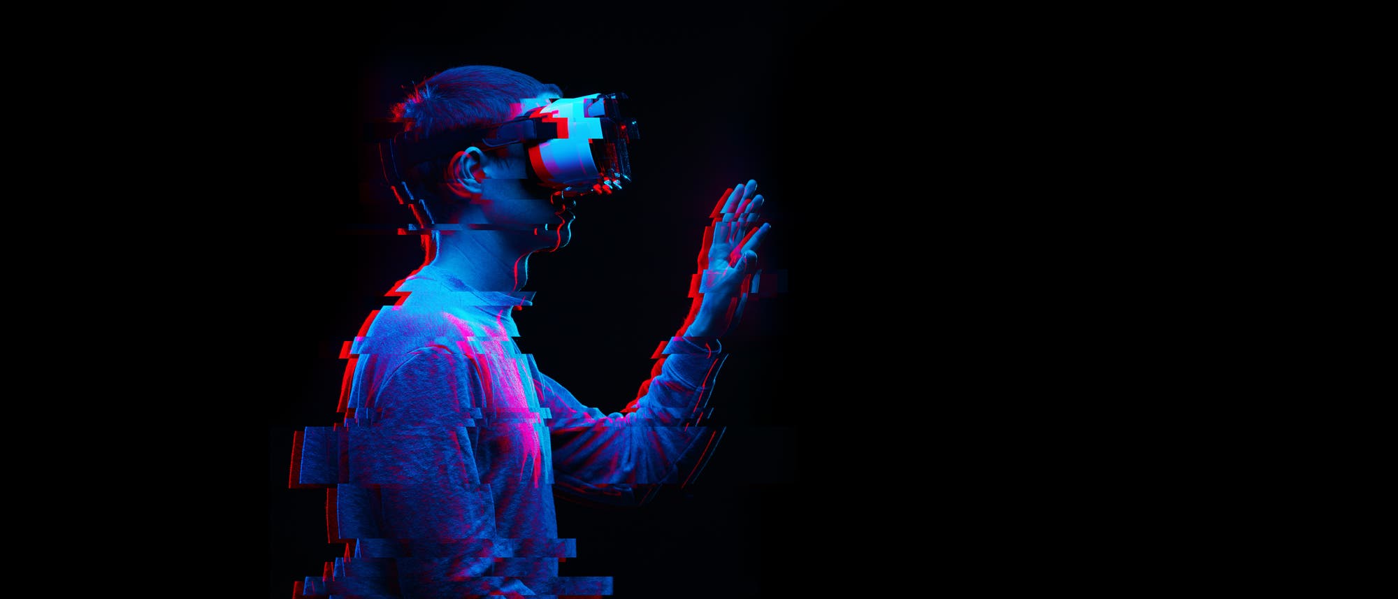 Eine Person tr&auml;gt eine Virtual-Reality-Brille, hat tastend eine Hand in die Luft gehoben und scheint in eine virtuelle Umgebung vertieft zu sein. Das Bild ist rot und blau verpixelt &uuml;berlagert, was die technische Atmosph&auml;re nachzeichnet.