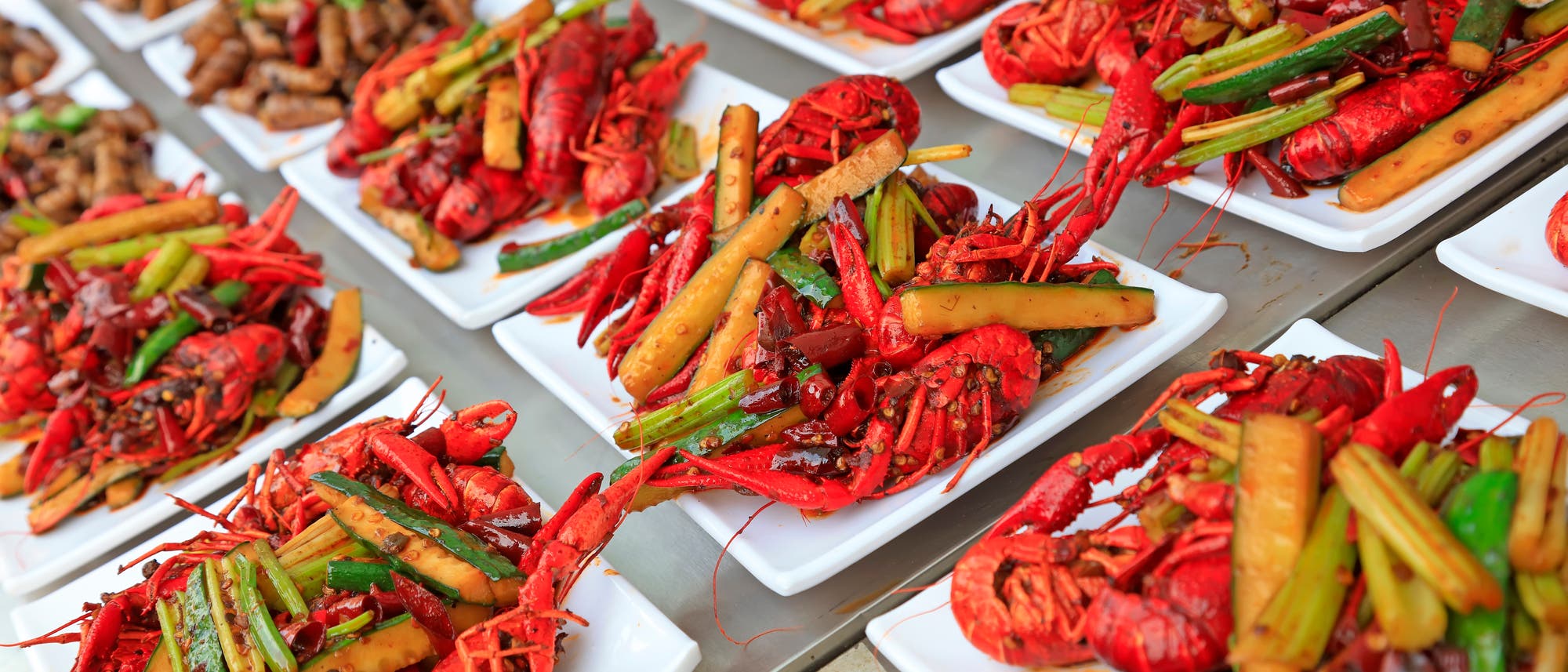 Eine Reihe von Tellern, die mit gekochten Flusskrebsen und Gemüse wie Frühlingszwiebeln und Gurken garniert sind. 