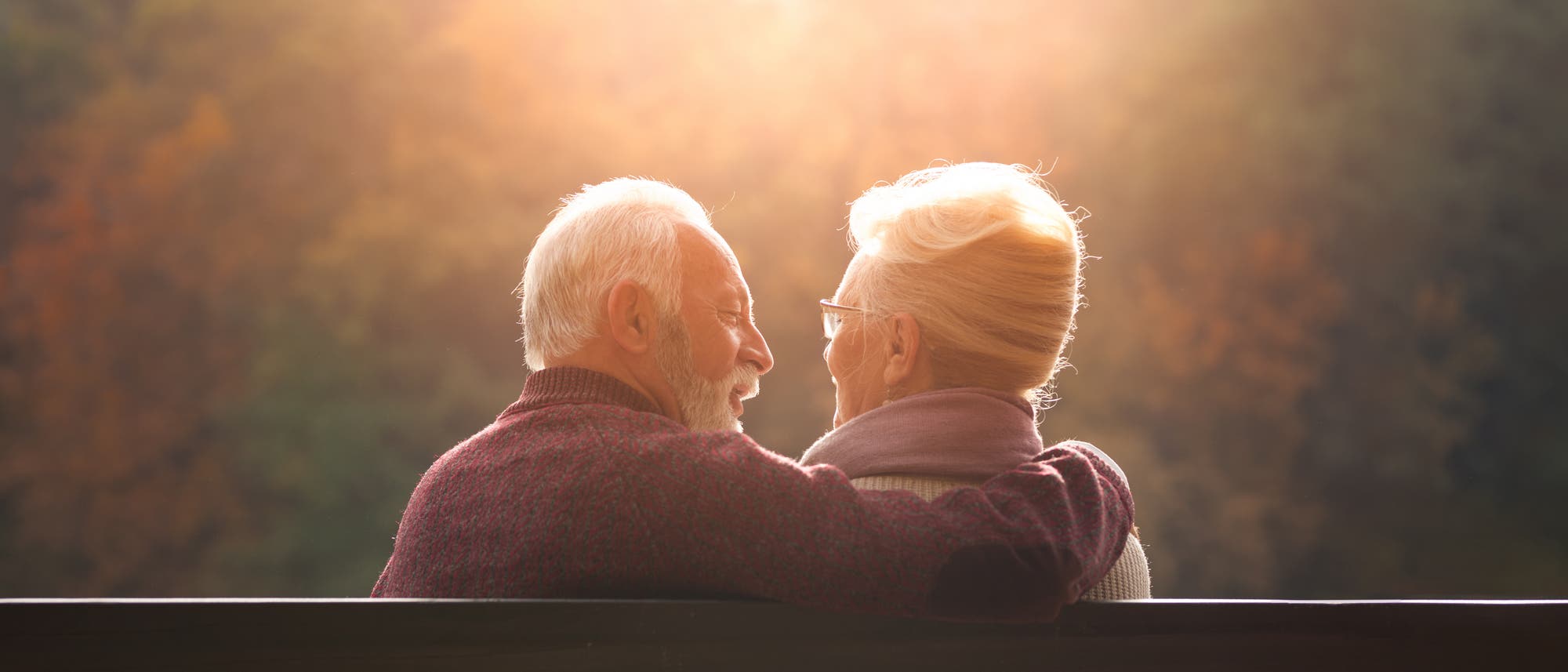 Ein älteres Paar sitzt auf einer Bank und schaut sich liebevoll an. Die Szene ist in warmes, goldenes Licht getaucht, das von der tiefstehenden Sonne im Hintergrund erzeugt wird. Im Hintergrund sind unscharfe Herbstbäume zu sehen, die eine ruhige und friedliche Atmosphäre schaffen. Ein älteres Paar sitzt auf einer Bank und schaut sich liebevoll an. Die Szene ist in warmes, goldenes Licht getaucht, das von der tiefstehenden Sonne im Hintergrund erzeugt wird. Im Hintergrund sind unscharfe Herbstbäume zu sehen, die eine ruhige und friedliche Atmosphäre schaffen.