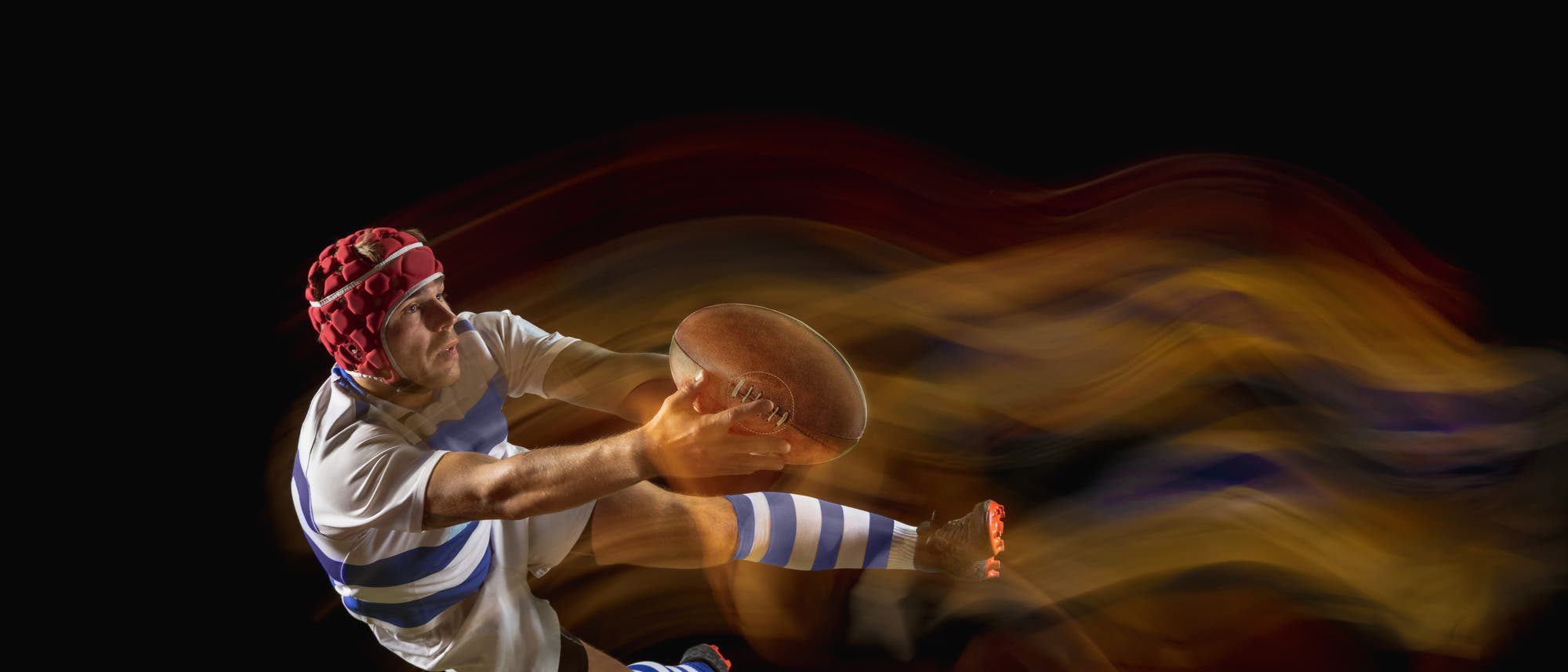 Ein Rugbyspieler in einem gestreiften Trikot und Helm springt dynamisch durch die Luft, w&auml;hrend er einen Rugbyball festh&auml;lt. 