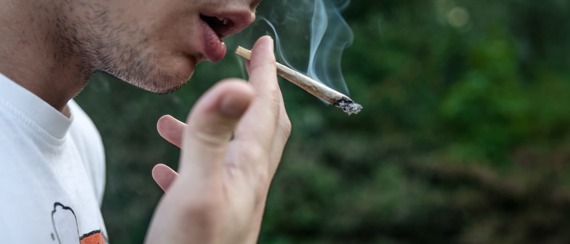 Eine Person h&auml;lt einen rauchenden Joint nahe am Mund. Der Hintergrund ist unscharf und zeigt gr&uuml;ne Vegetation. Die Person tr&auml;gt ein wei&szlig;es T-Shirt mit einem teilweise sichtbaren Aufdruck. Rauch steigt vom Joint auf.