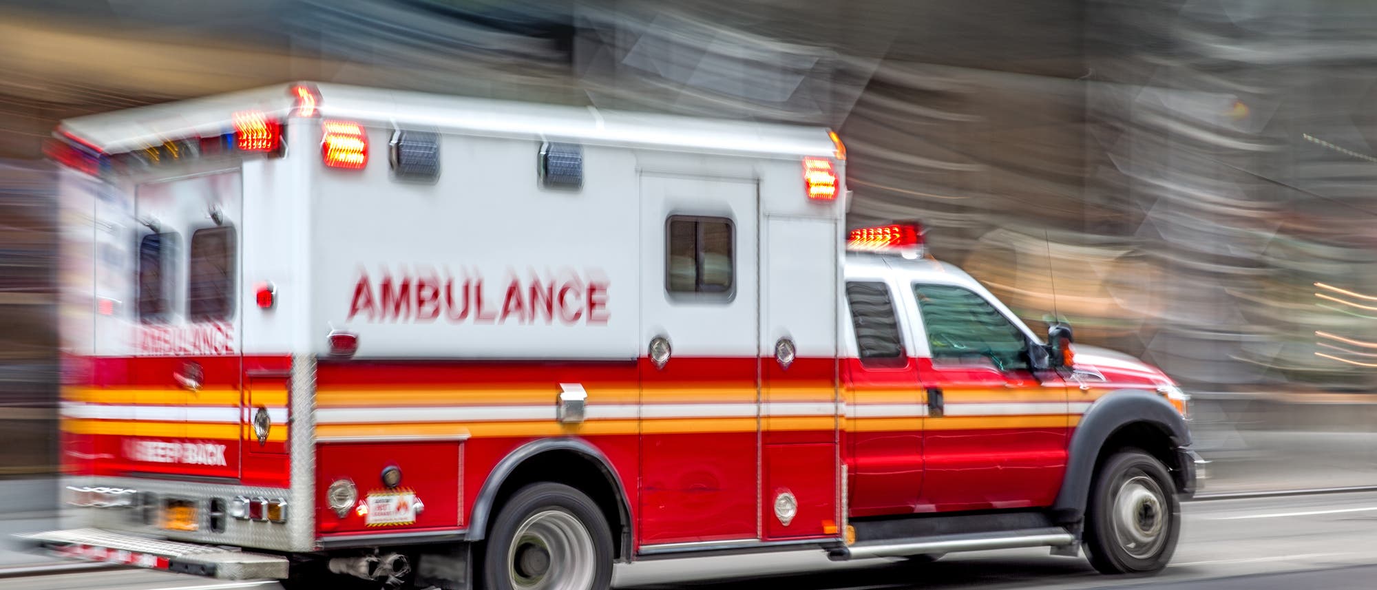 Ein rotes und weißes Krankenwagenfahrzeug mit eingeschaltetem Blaulicht fährt schnell eine Straße entlang. Auf der Seite des Fahrzeugs steht „AMBULANCE“. Die Umgebung ist verschwommen, was die Bewegung des Fahrzeugs betont.