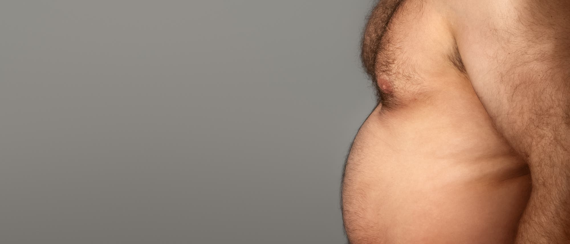 Ein Foto zeigt den seitlichen Oberkörper eines Menschen mit freiem Oberkörper vor einem grauen Hintergrund. Der Fokus liegt auf dem Bauchbereich.