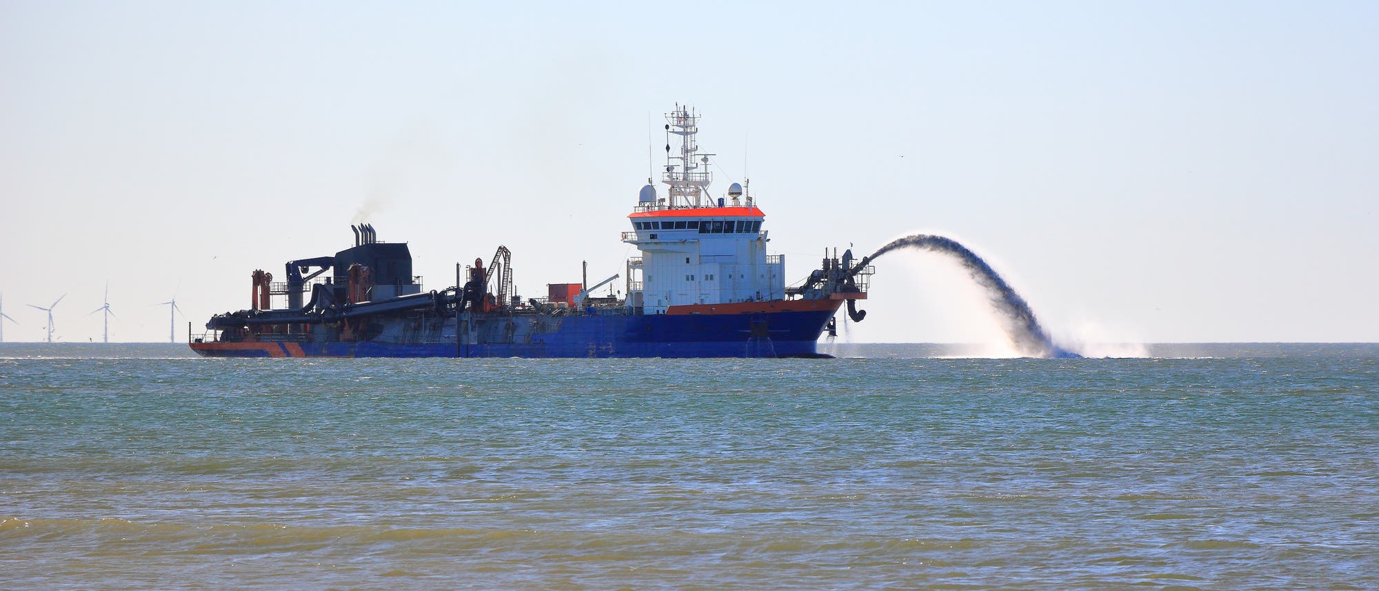 Ein gro&szlig;es blaues Schiff, das als Saugbagger fungiert, befindet sich auf dem Meer. Es spr&uuml;ht Wasser und Sand in einem Bogen zur&uuml;ck ins Meer. Im Hintergrund sind Windkraftanlagen am Horizont zu sehen. Der Himmel ist klar und blau.