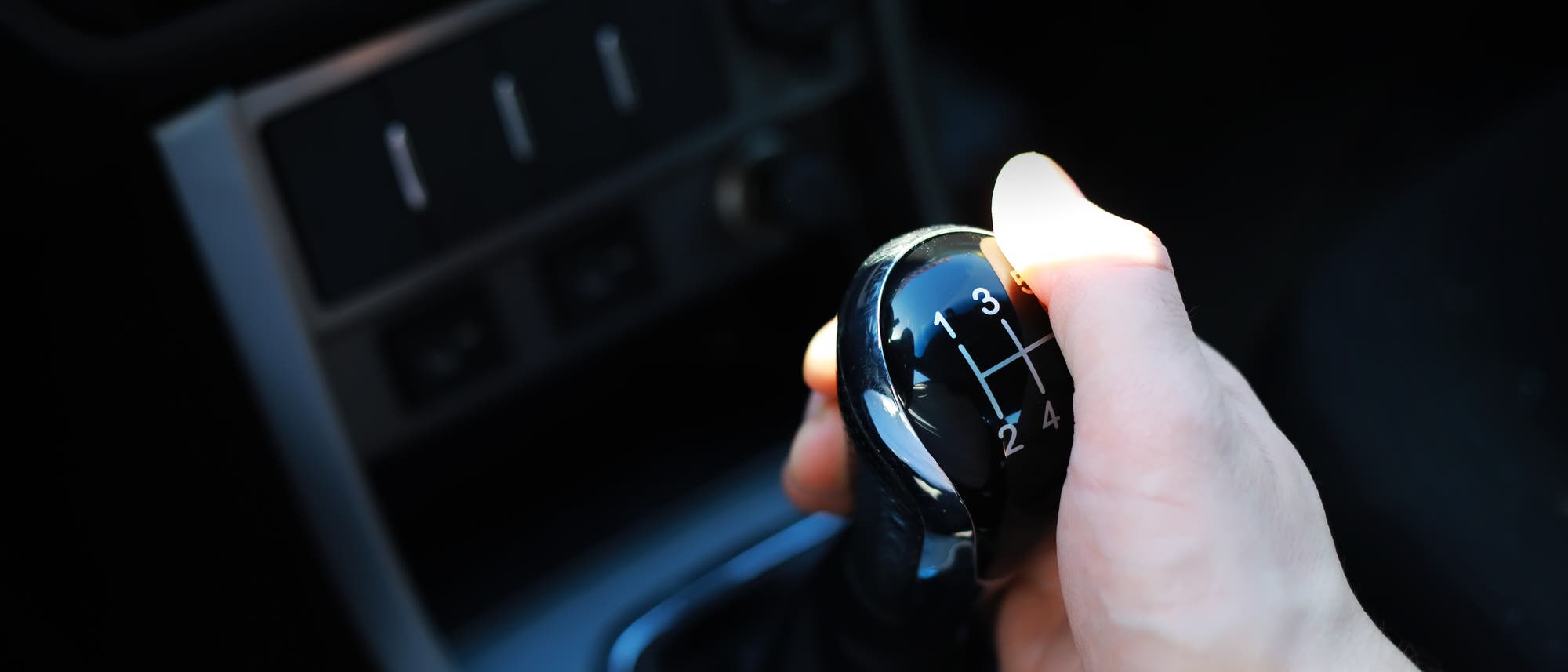 Eine Hand hält den Schalthebel eines Autos, der die Gänge 1 bis 5 und den Rückwärtsgang anzeigt. Der Schalthebel befindet sich in einem Fahrzeugcockpit mit sichtbaren Bedienelementen im Hintergrund.