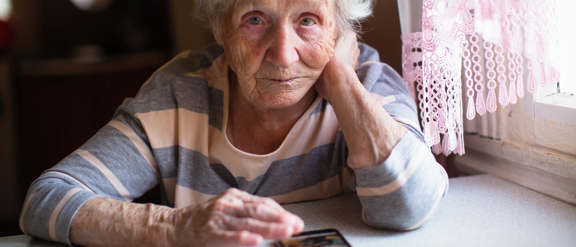 Eine ältere Person sitzt an einem Tisch und schaut nachdenklich in die Kamera. Die Person trägt ein gestreiftes Oberteil und stützt den Kopf auf eine Hand. Auf dem Tisch liegt ein Smartphone. Rechts im Bild hängt ein rosa Vorhang am Fenster. Die Szene wirkt ruhig und nachdenklich.