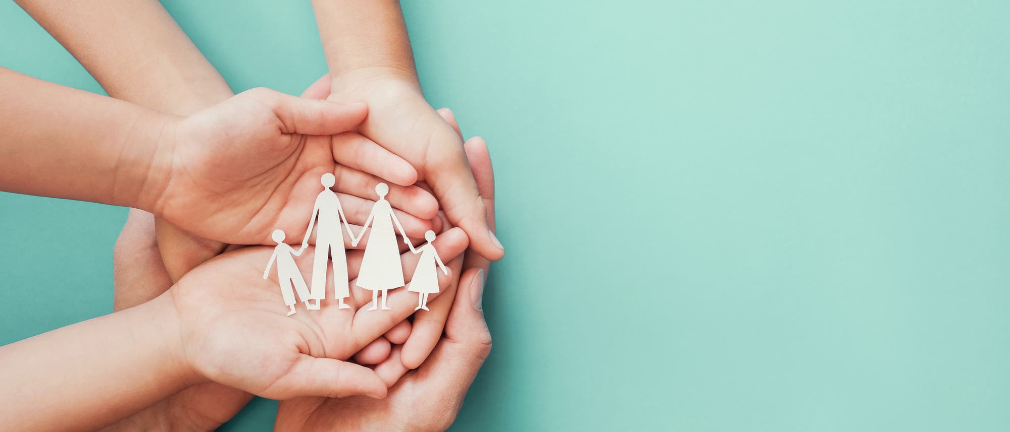 Hände von Erwachsenen und Kindern halten gemeinsam ausgeschnittene Papierfiguren einer Familie auf einem türkisfarbenen Hintergrund. Die Figuren stellen zwei Erwachsene und zwei Kinder dar, die sich an den Händen halten. Das Bild symbolisiert Schutz, Fürsorge und Zusammenhalt innerhalb einer Familie. Hände von Erwachsenen und Kindern halten gemeinsam ausgeschnittene Papierfiguren einer Familie auf einem türkisfarbenen Hintergrund. Die Figuren stellen zwei Erwachsene und zwei Kinder dar, die sich an den Händen halten. Das Bild symbolisiert Schutz, Fürsorge und Zusammenhalt innerhalb einer Familie.