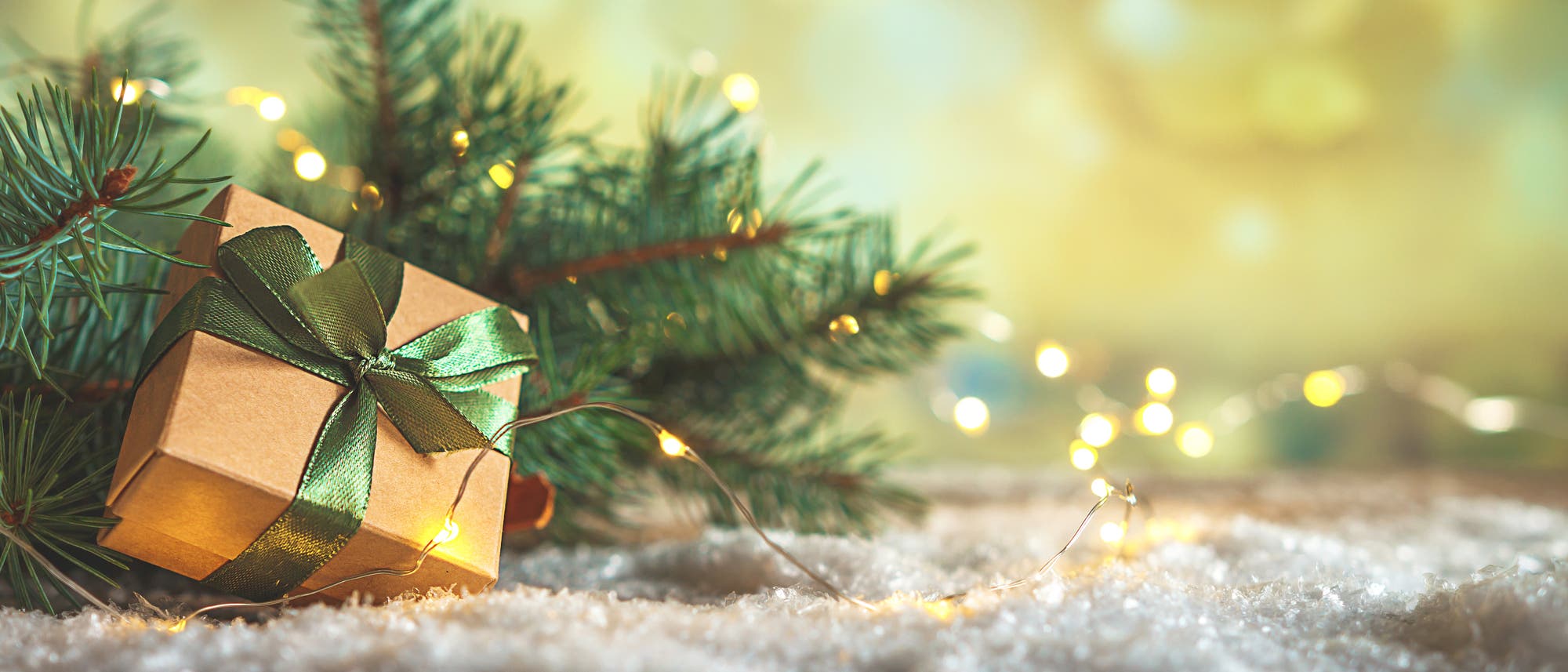 Eine Geschenkbox in braunem Papier ist mit einer gr&uuml;nen Schleife verziert. Im Hintergrund sind Tannenzweige, Glitzer und eine Lichterkette zu sehen. 