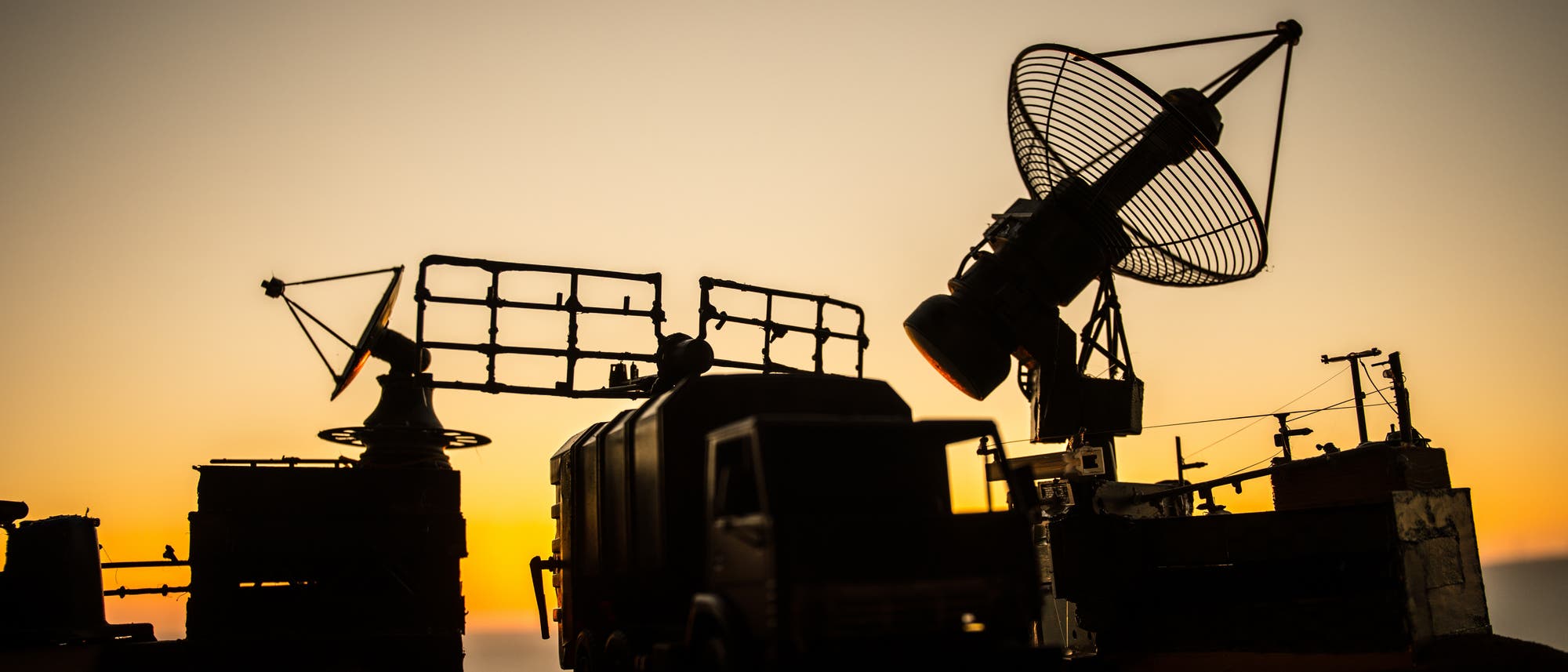 Silhouette einer militärischen Radaranlage bei Sonnenuntergang. Im Vordergrund ist ein Lastwagen zu sehen, umgeben von mehreren großen Radarantennen. Die Antennen sind auf Plattformen montiert und zeigen in verschiedene Richtungen. Der Himmel im Hintergrund ist in Gelb- und Orangetönen gefärbt.