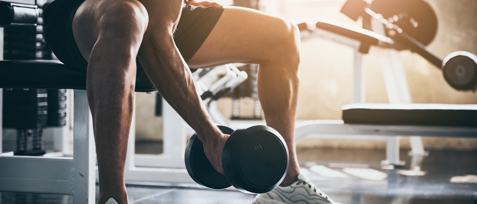 Ein Mann hebt im Fitnessstudio ein Gewicht an. Die Sonne scheint auf seinen muskulösen Oberarm 